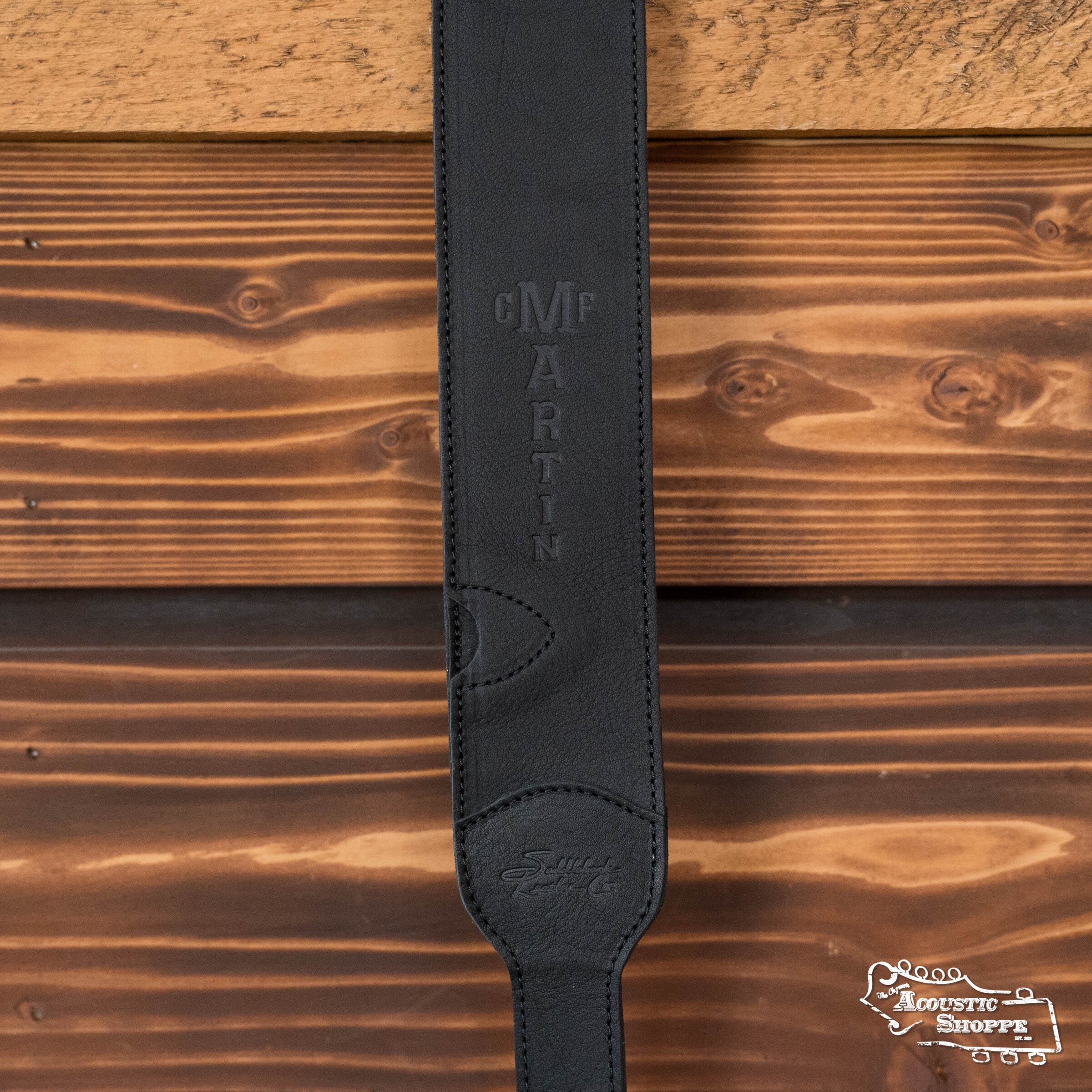 The LUXE By Martin Black Leather Guitar Strap from Martin & Co. hangs against a wooden wall, featuring an embossed logo. The Acoustic Shoppe logo appears in white at the bottom right corner.