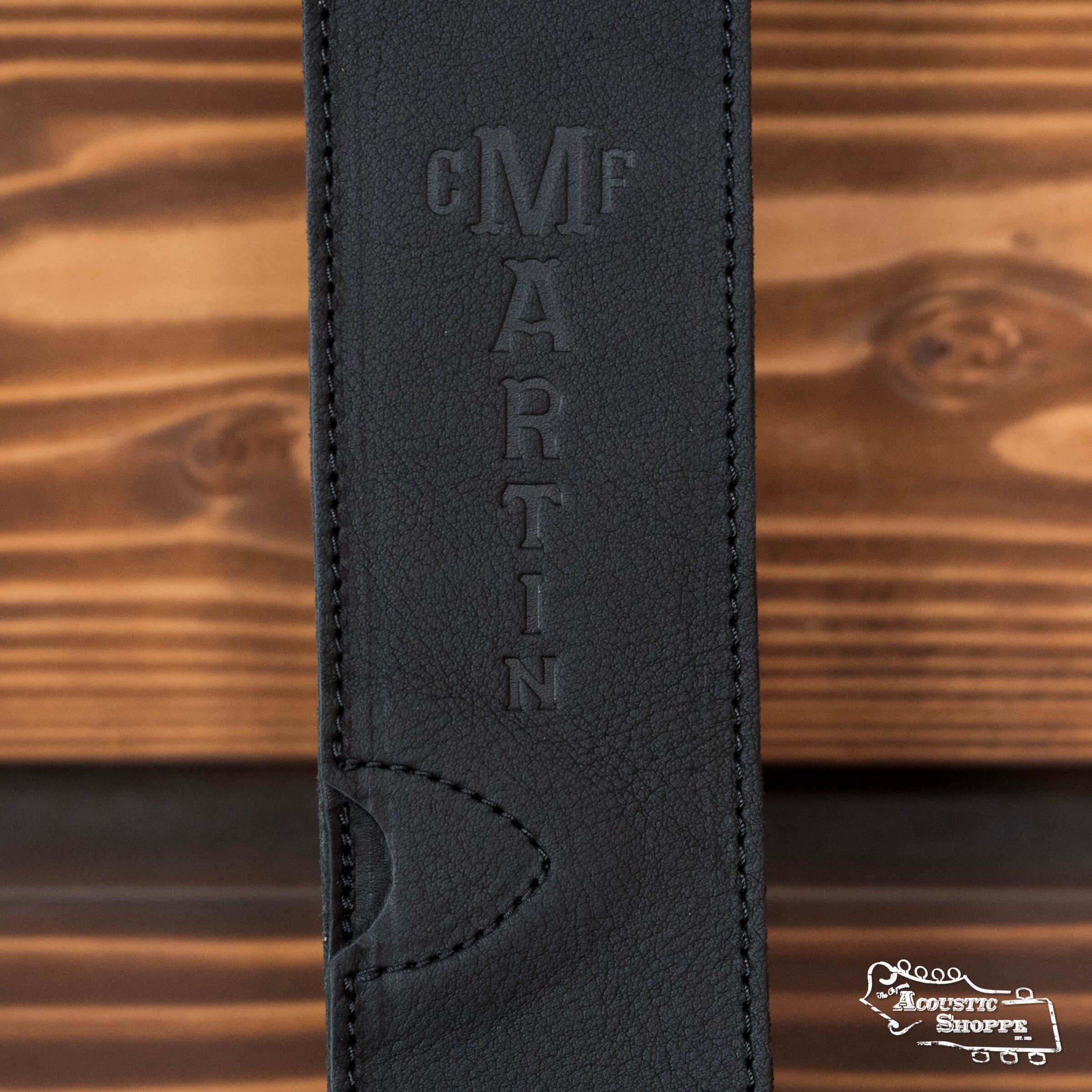 Close-up of the LUXE By Martin Black Leather Guitar Strap by Martin & Co., featuring full grain black leather and a vertical Martin logo. The strap rests on wood beside The Acoustic Shoppe logo, adding premium style to your setup.