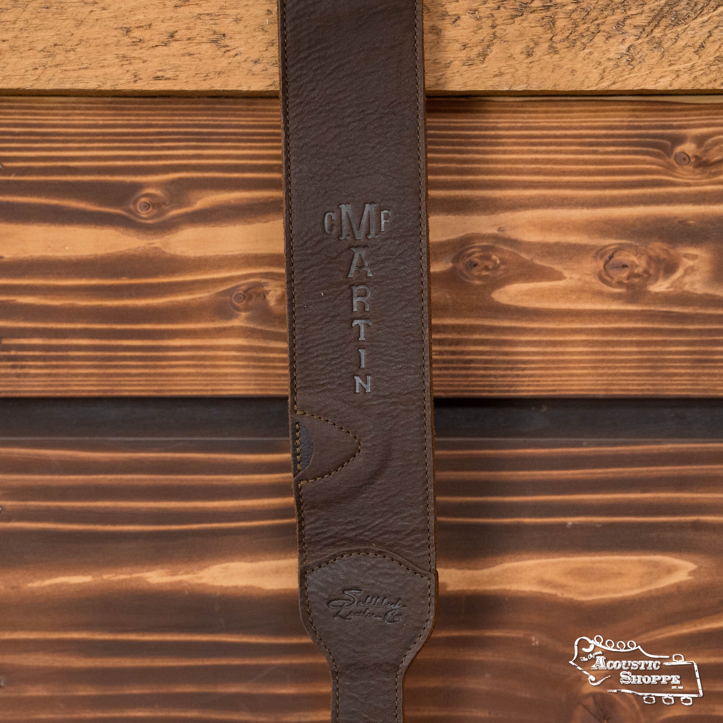 A close-up of a LUXE By Martin Dark Brown Leather Guitar Strap from Martin & Co. is shown against a wooden background, with The Acoustic Shoppe logo in the bottom right corner.