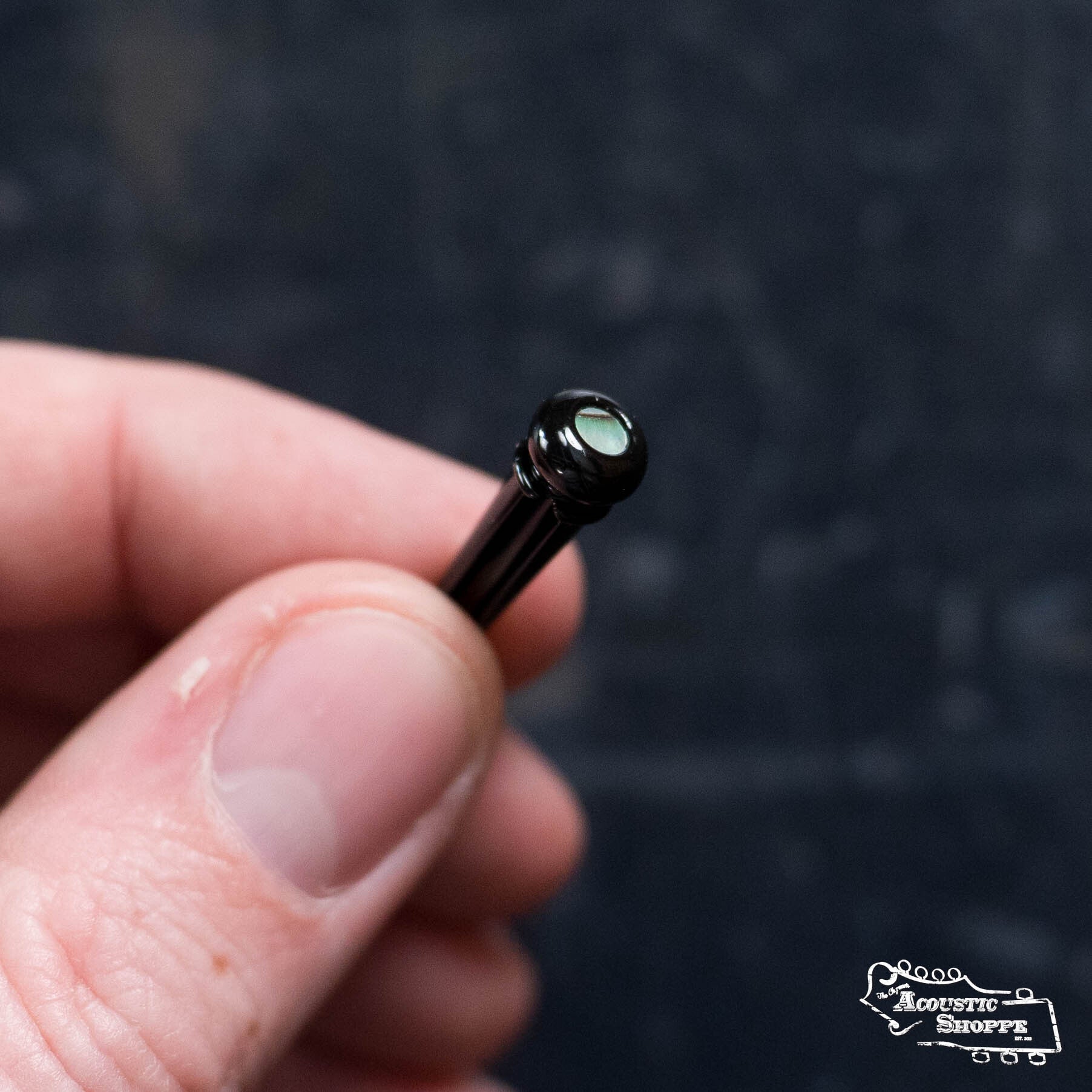 A hand holds a Martin & Co. LUXE Pack of 6 Bridge Pins in gloss black with pearl inlay, set against a dark, blurred background. The Acoustic Shoppe logo appears in the bottom right corner.
