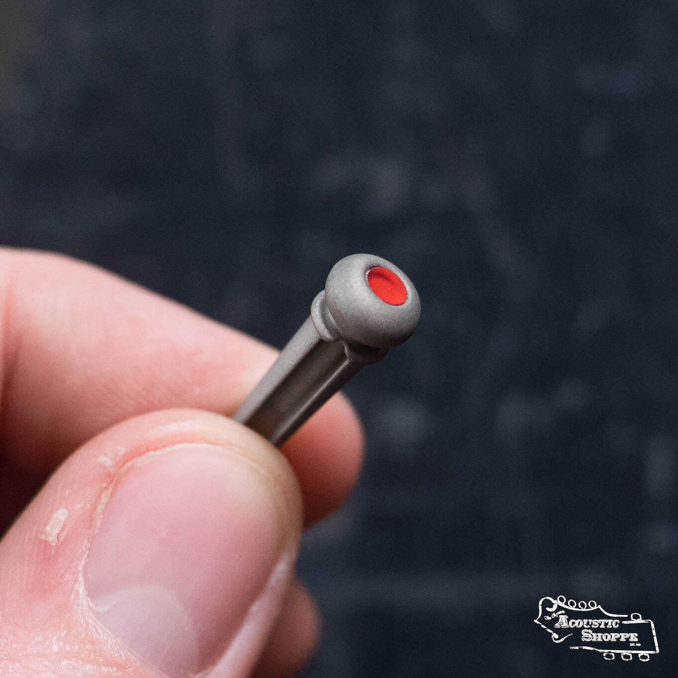 A hand holds a small, gray LUXE By Martin Turkish Emery bridge pin with a red dot, against a dark background. The Acoustic Shoppe logo at the bottom right highlights these premium Martin & Co. accessories, sold in packs of six.