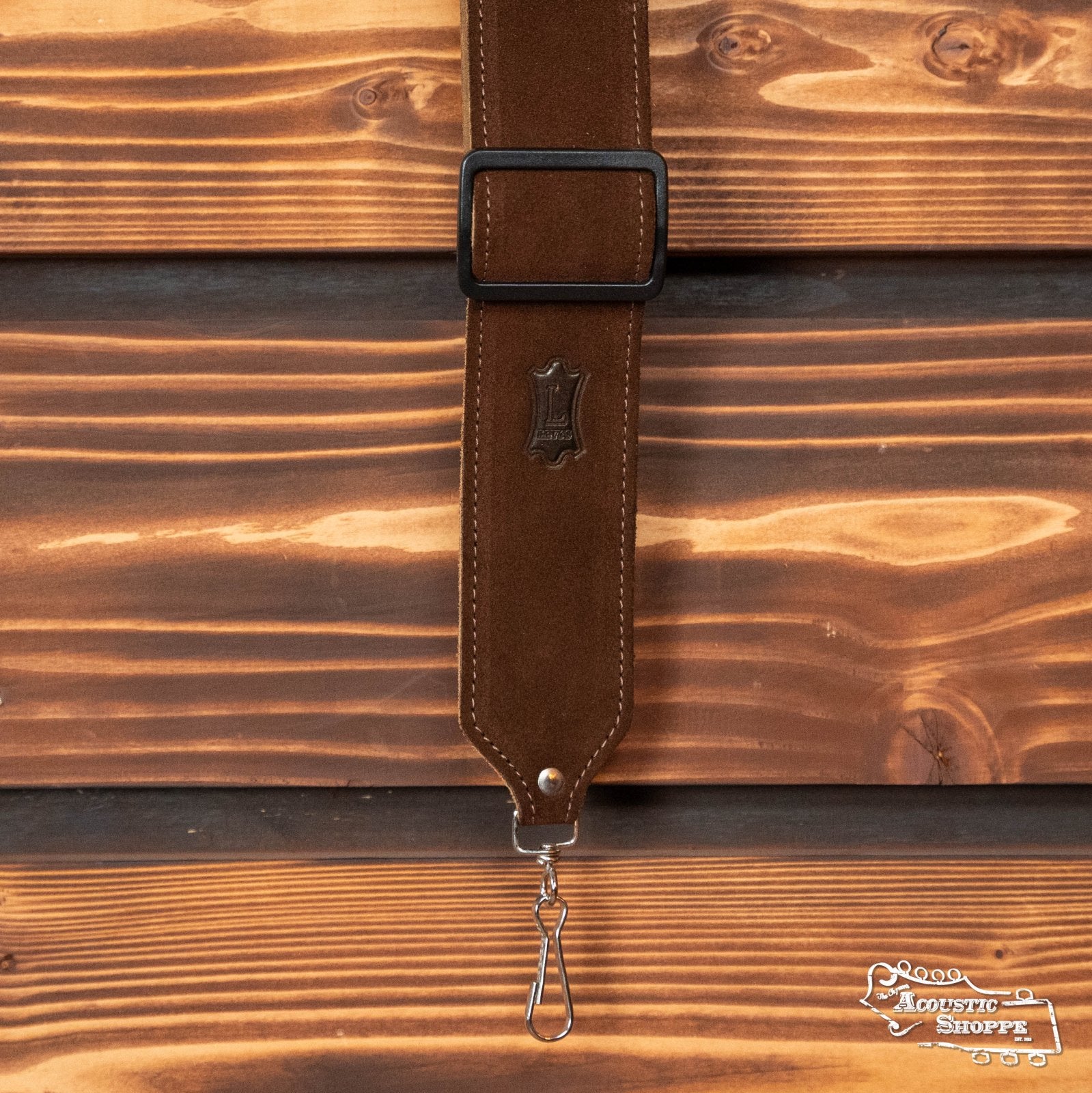 The Levys Leather Banjo Strap (M9S-BRN) by Levys Leather, in brown suede, hangs against a wooden wall. It features a black adjustment buckle, metal clips, visible stitching, and a small embossed logo.