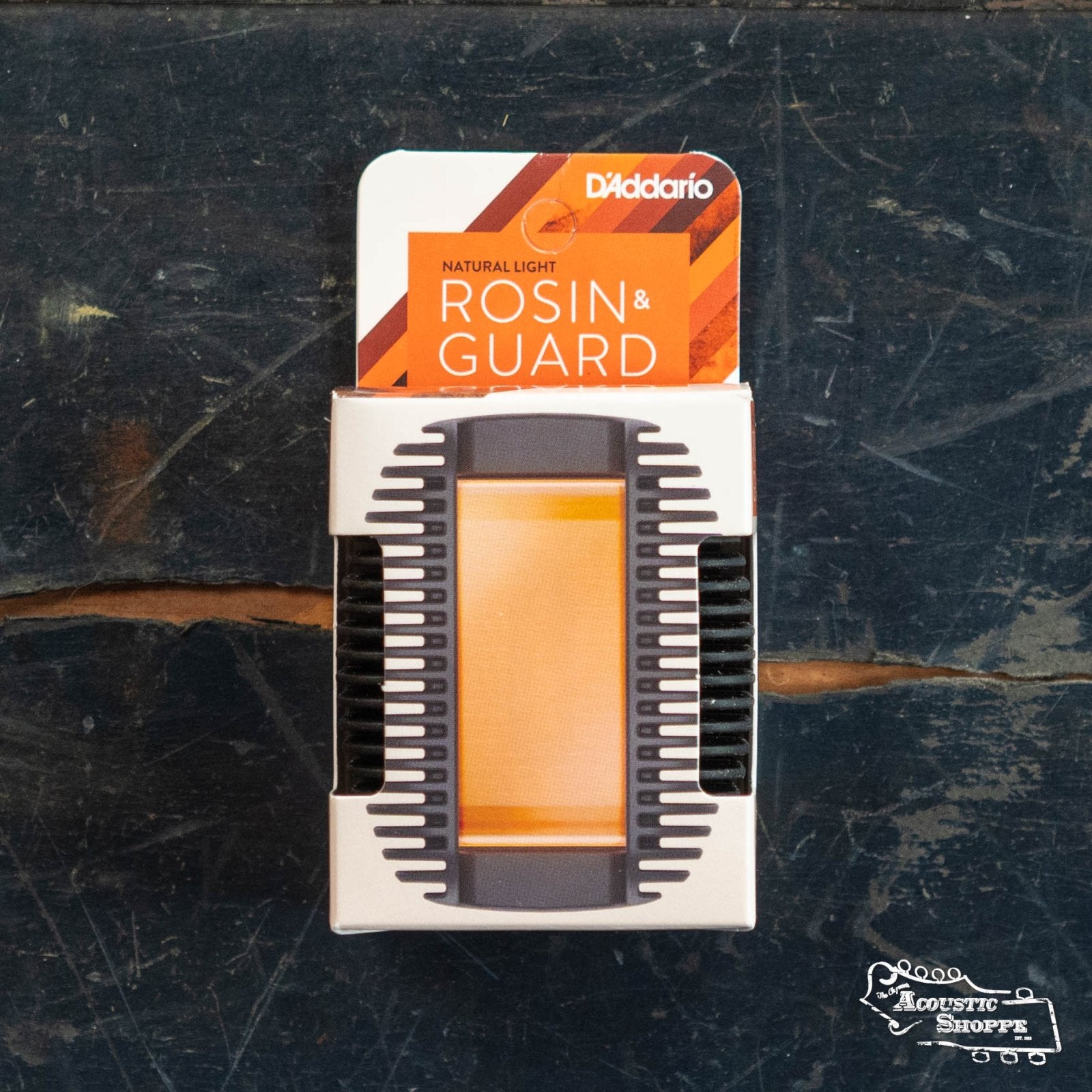 A box of DAddario DRGBK-VR200 Rosin Guard with Natural Light Rosin for acoustic instruments rests on a worn dark wooden surface. The white and orange packaging has bold text and a clear window showing the rosin inside.
