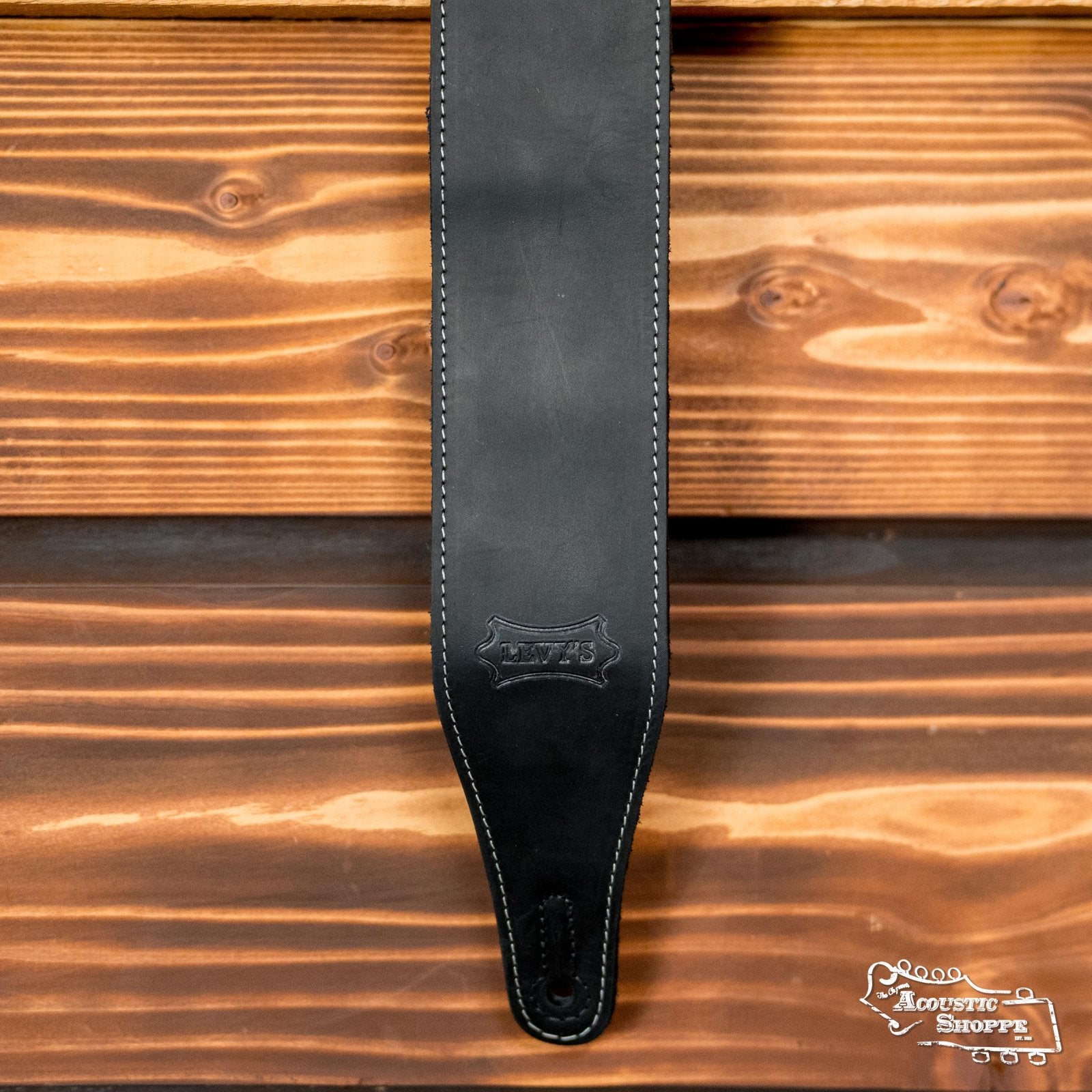 A Levys Leather 2.5 Pull-Up Butter Leather Guitar Strap in black (M17BAS-BLK) with white stitching, stamped logo, and pre-cut end hole hangs against a wooden background, ready for easy guitar attachment.