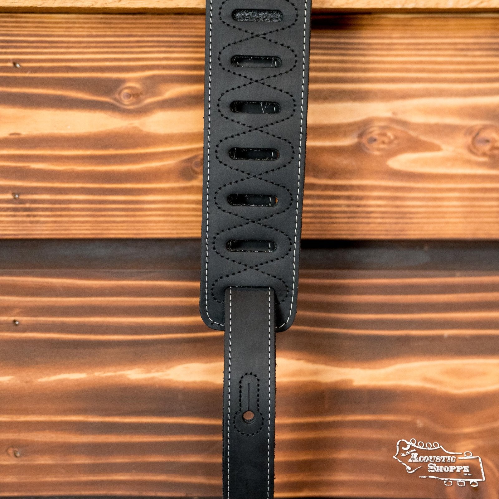 A Levys 2.5 Pull-Up Butter Leather Guitar Strap in black (M17BAS-BLK) hangs in front of a rustic wooden wall with The Acoustic Shoppe logo in the bottom right corner.