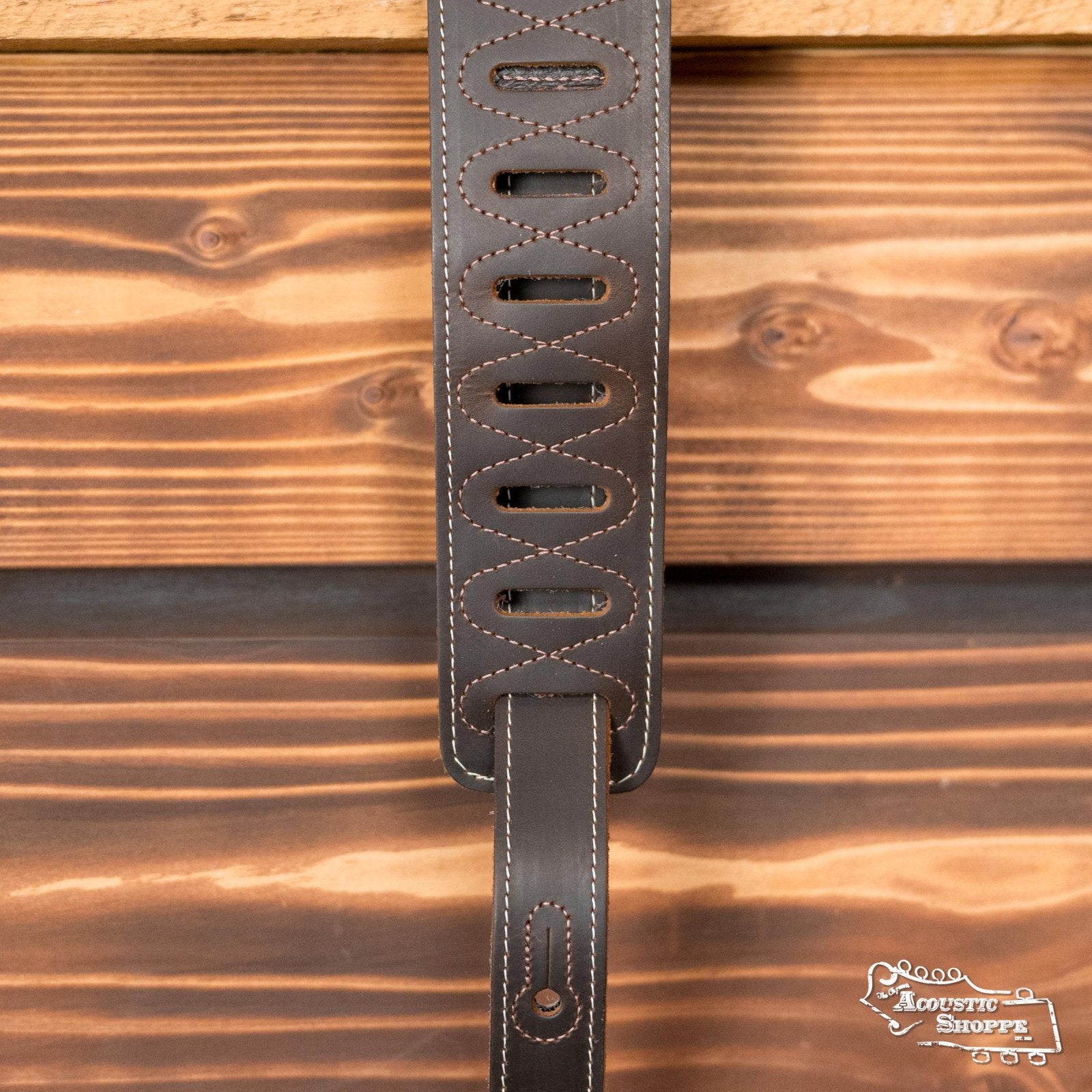 A Levy's Leather 2.5" Pull-Up Butter Leather Guitar Strap in brown (M17BAS-DBR) with oval cutouts and white stitching hangs on a wood wall. The Acoustic Shoppe logo marks this trusted Eastman dealer for acoustic instruments.