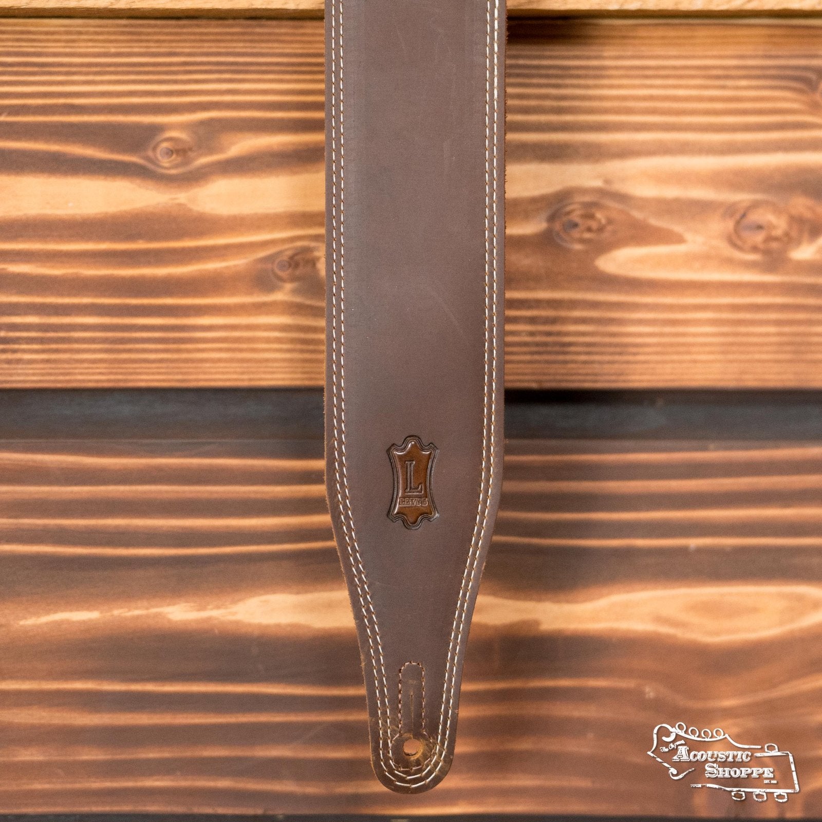 The Levys Leather M17BDS-BRN Like Butter Double Stitch Brown Guitar Strap, featuring an embossed āLā emblem and adjustable design, hangs against wood with The Acoustic Shoppe logo in the bottom right corner.