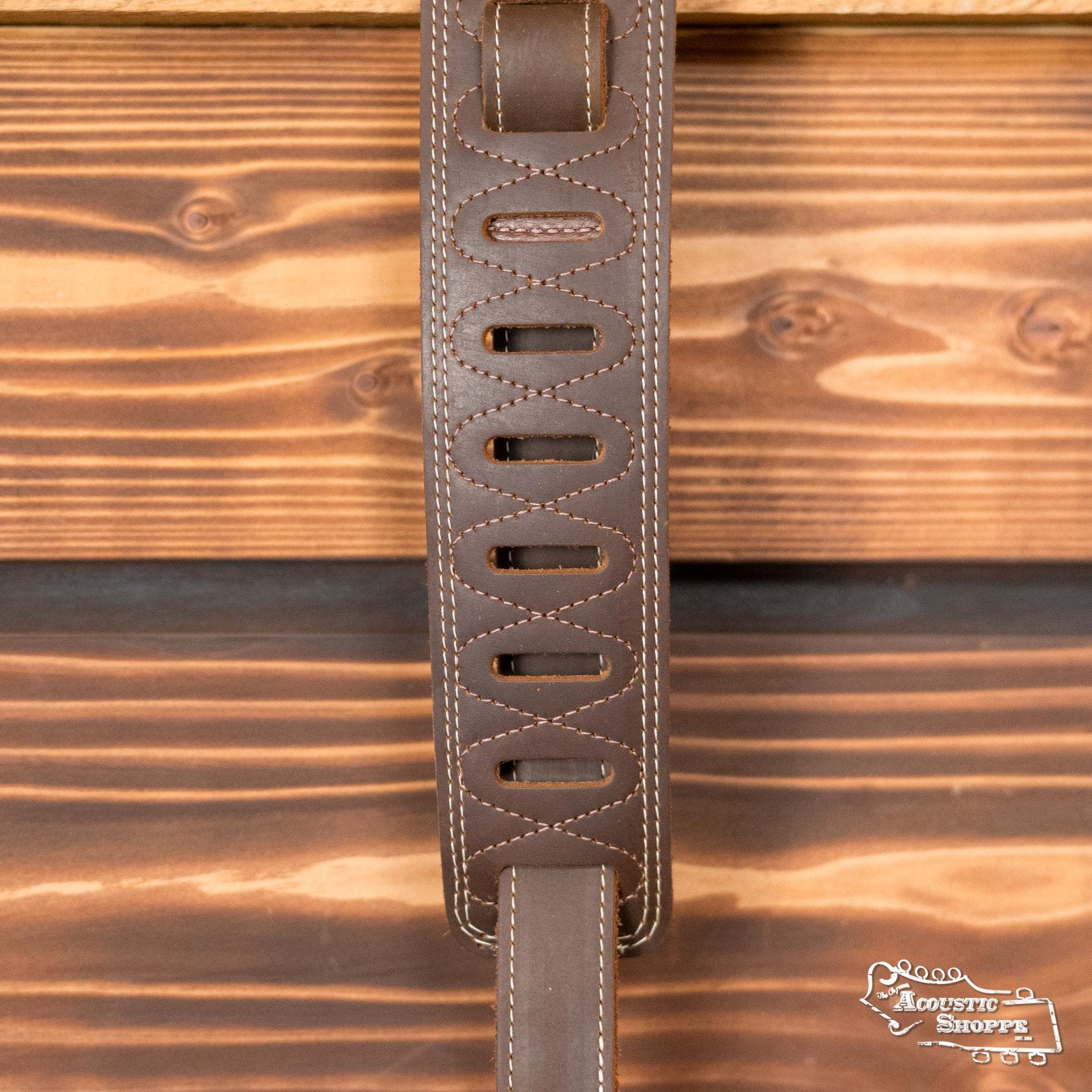The Levys Leather M17BDS-BRN Like Butter Double Stitch Leather Guitar Strap in brown, with decorative cutouts and white stitching, hangs against a wooden background. The Acoustic Shoppe logo appears in the lower right corner.