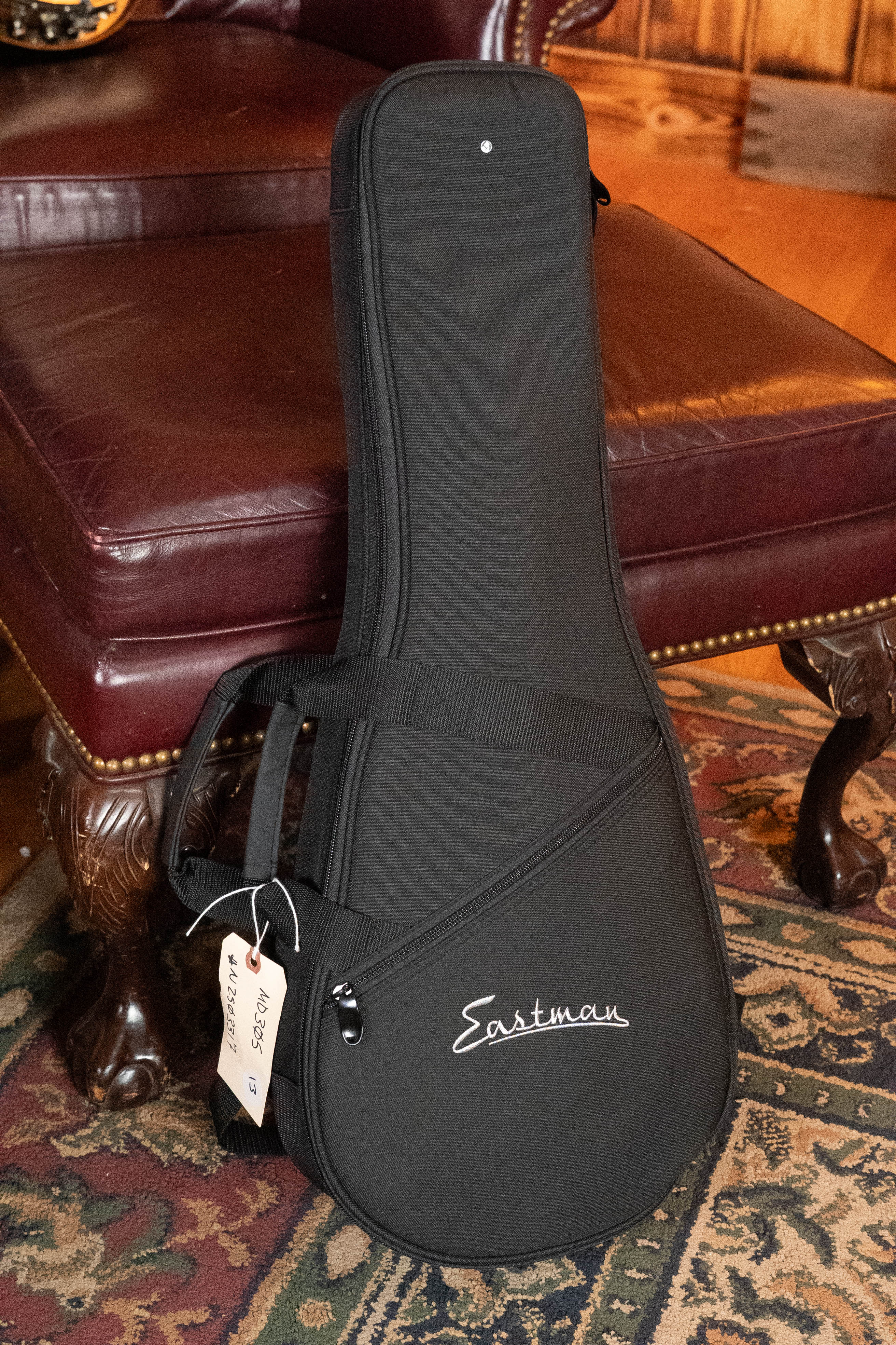 An Eastman Guitars MD305 Spruce/Maple A-Style hand-carved mandolin (#3317) in a black case with shoulder strap and tag stands upright on a patterned rug before a maroon leather chair with ornate wood legs.
