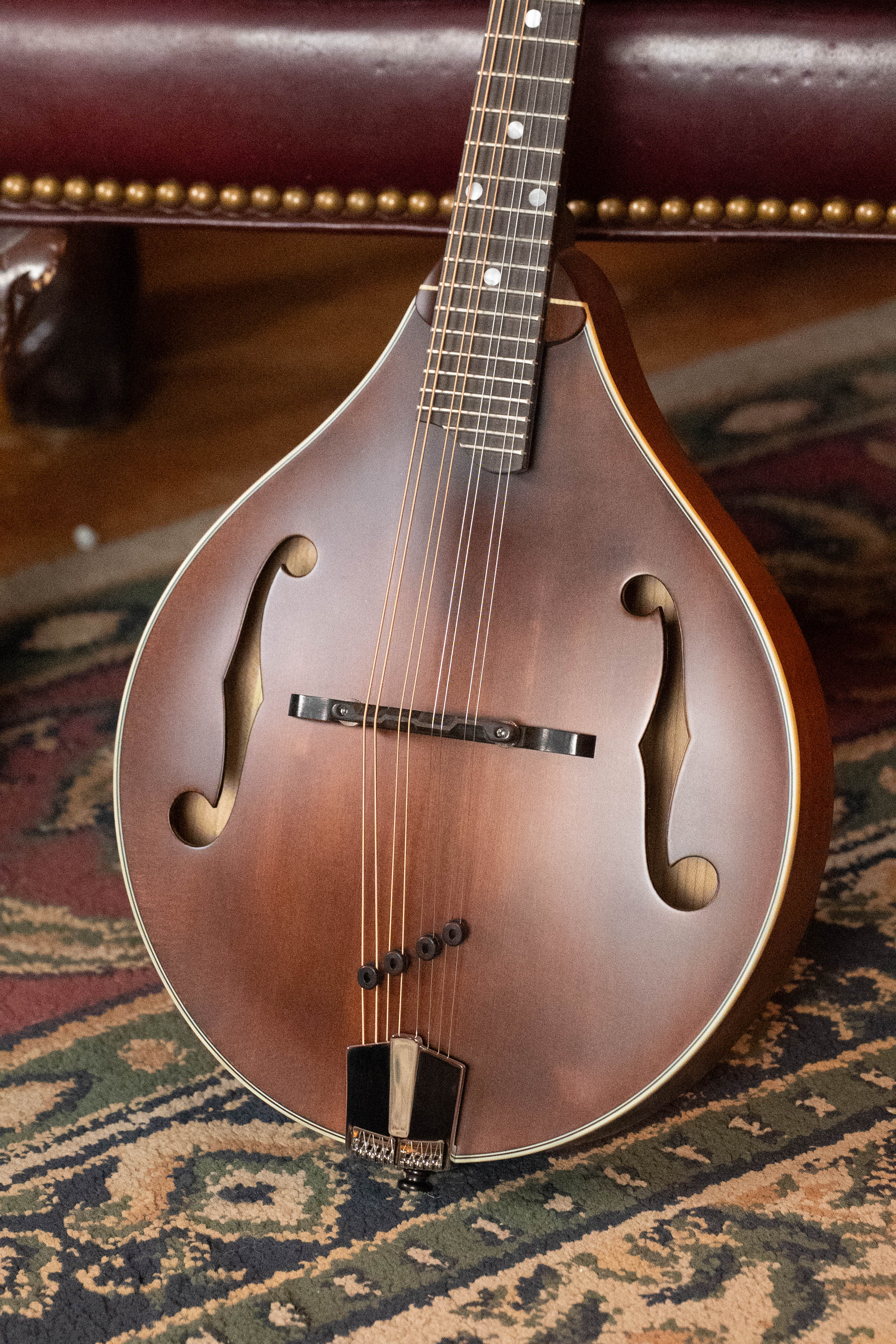 An Eastman Guitars MD305 Spruce/Maple A-Style Hand-Carved Mandolin (#3317) rests against a patterned rug and a burgundy leather chair with brass nailhead trim.