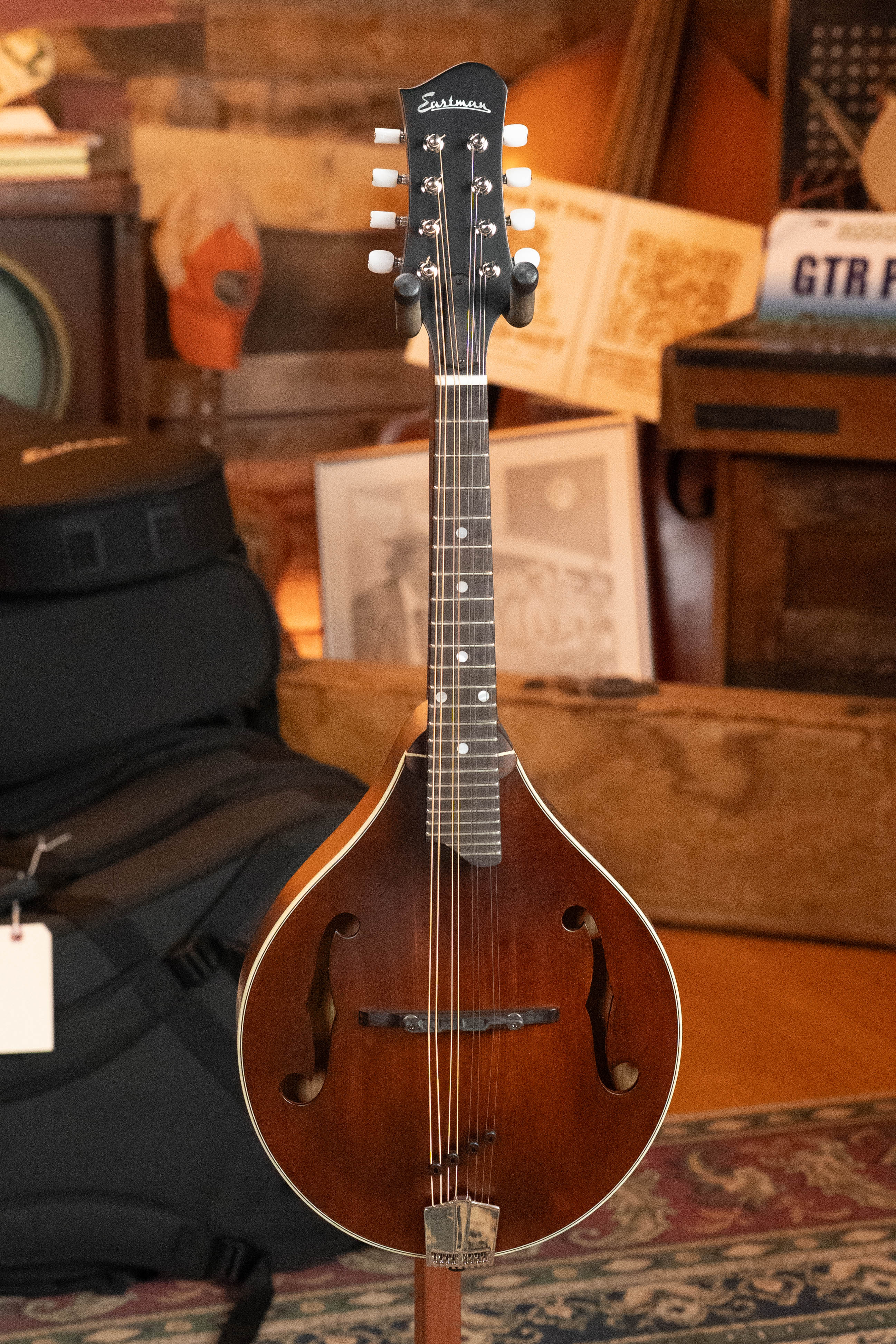 An Eastman Guitars MD305 Spruce/Maple A-Style Hand-Carved Mandolin (#3317) with a solid spruce top stands upright in a cozy, wood-paneled room filled with musical gear, framed art, and warm amber lighting.