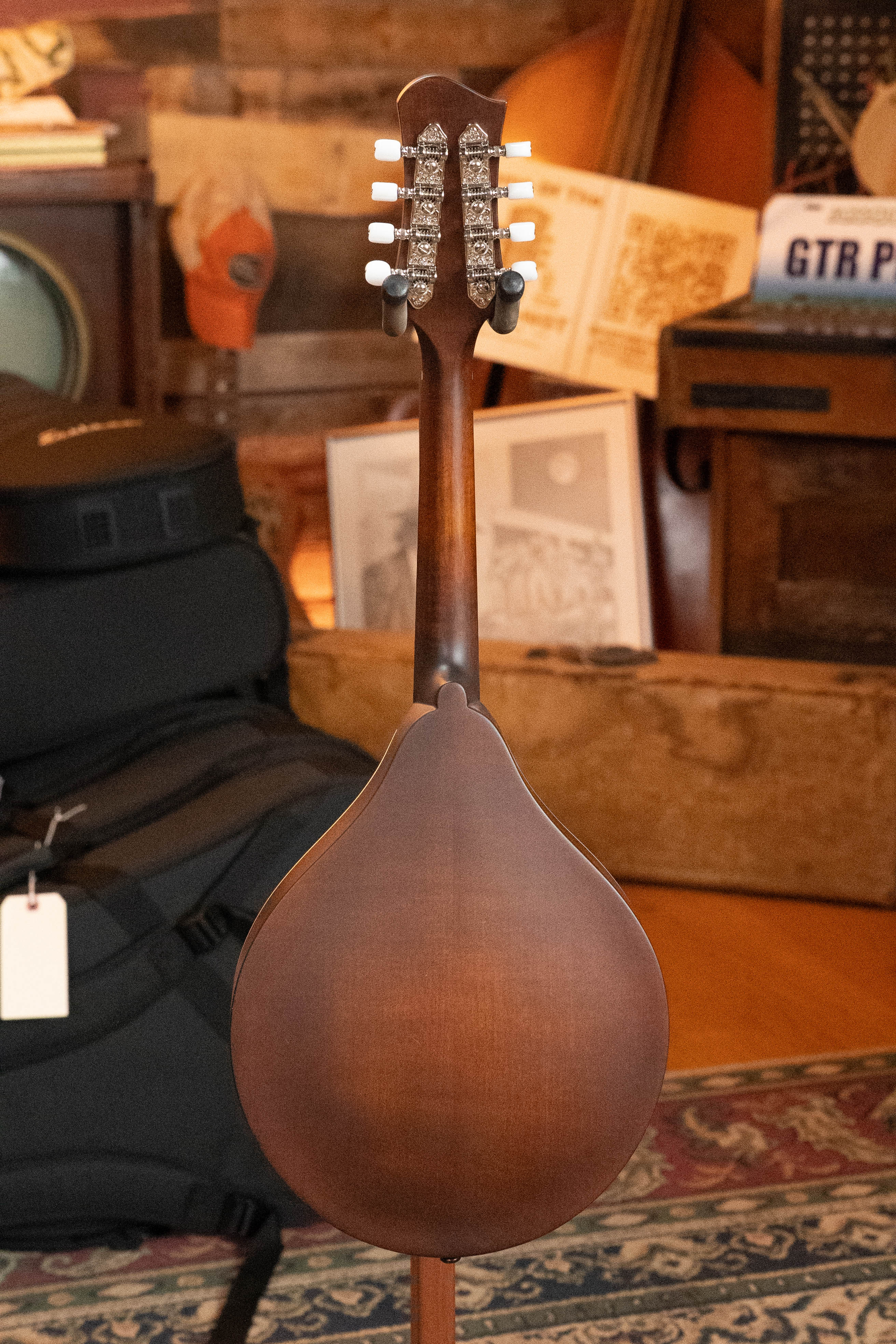 The Eastman Guitars MD305 Spruce/Maple A-Style Hand-Carved Mandolin #3317 is shown with its back facing the camera, set in a cozy room featuring musical instruments, a guitar case, and framed artwork in the background.
