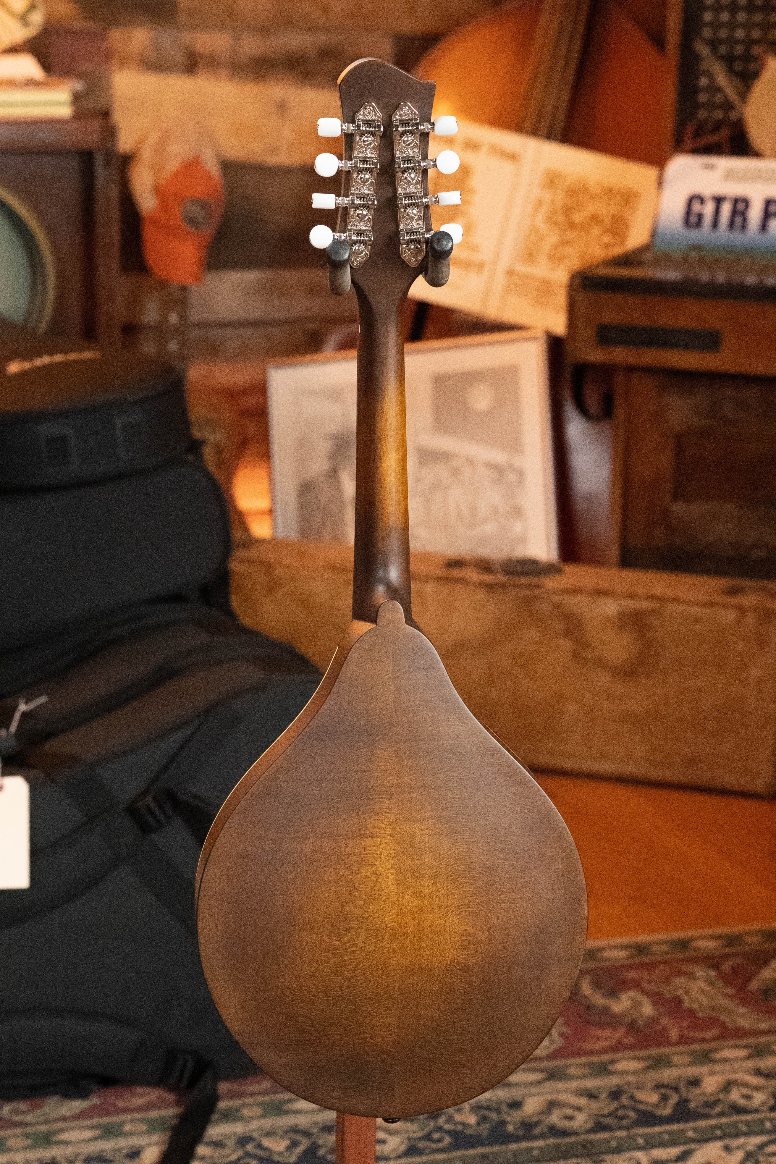 The Eastman Guitars MD305 Spruce/Maple A-Style Hand-Carved Mandolin #3596 is shown from the back, highlighting its wooden body and tuning pegs, with music gear, framed photos, and a patterned rug in the background.