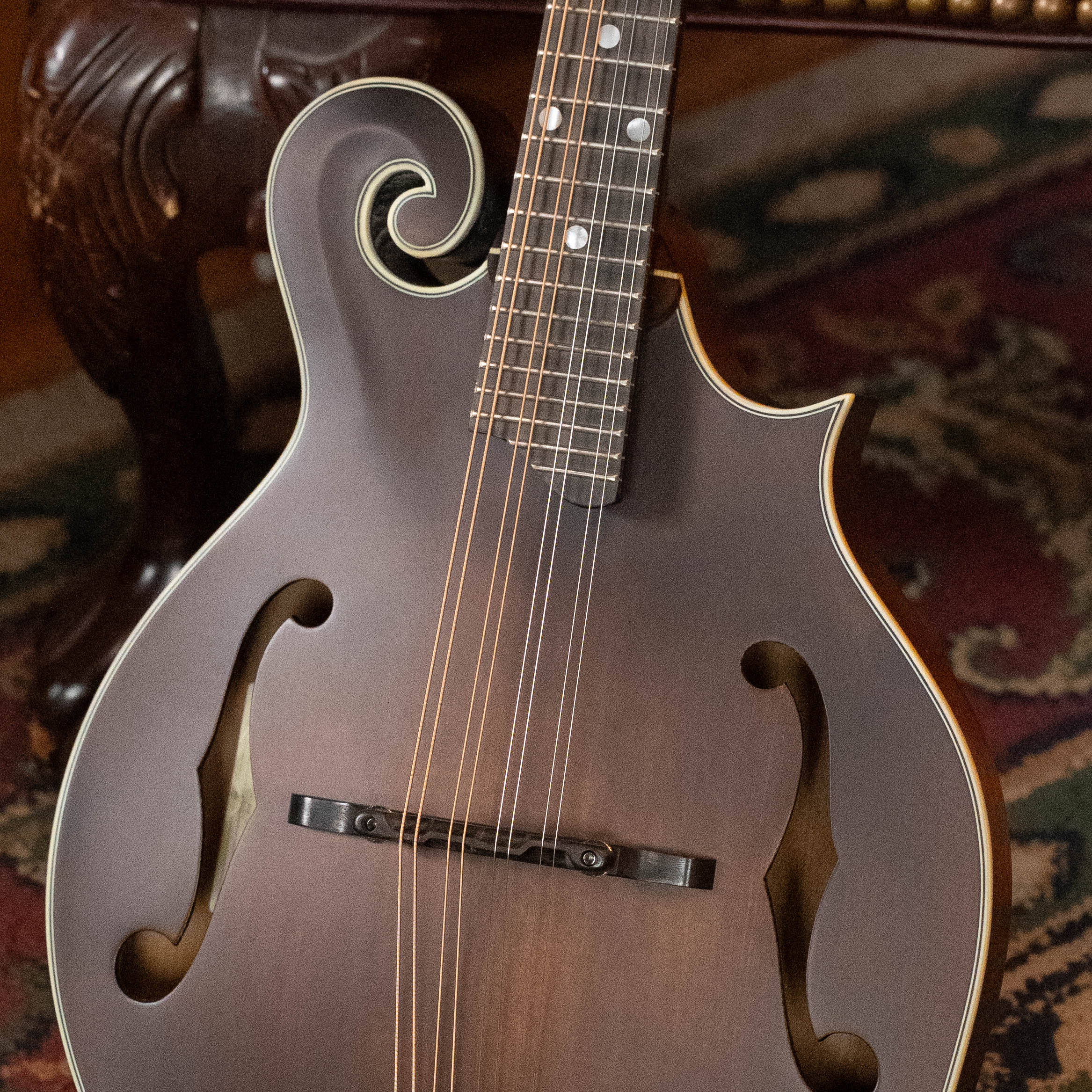 A close-up of an Eastman Guitars MD315 F-Style Hand-Carved Mandolin #0917 with f-holes and eight strings, resting on a patterned rug near ornately carved wooden furniture.