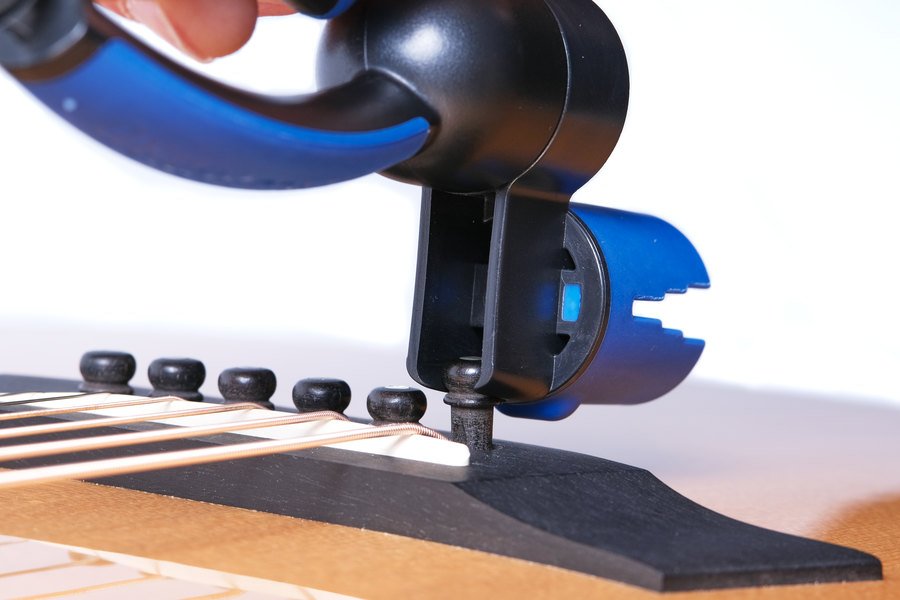 A close-up of a person using the Music Nomad GRIP ONE String Winder, Cutter, Puller (MN223) to remove a bridge pin from an acoustic guitar, with strings and the bridge clearly visible.