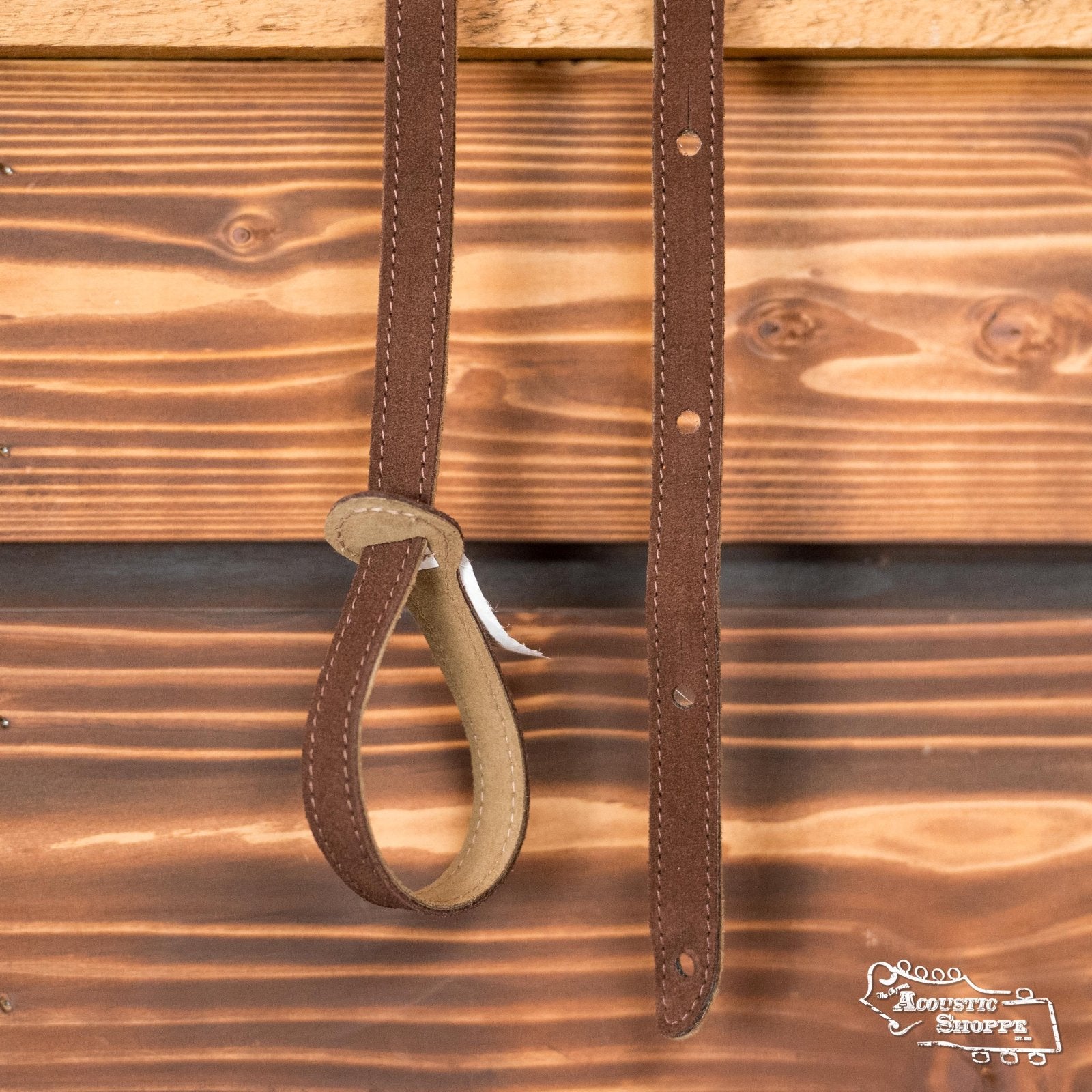 Two Levys Leather MS19-BRN 3/4 wide hand-brushed brown suede mandolin straps with holes hang against wood; one forms a loop at the bottom. The Acoustic Shoppe logo is in the bottom right corner.