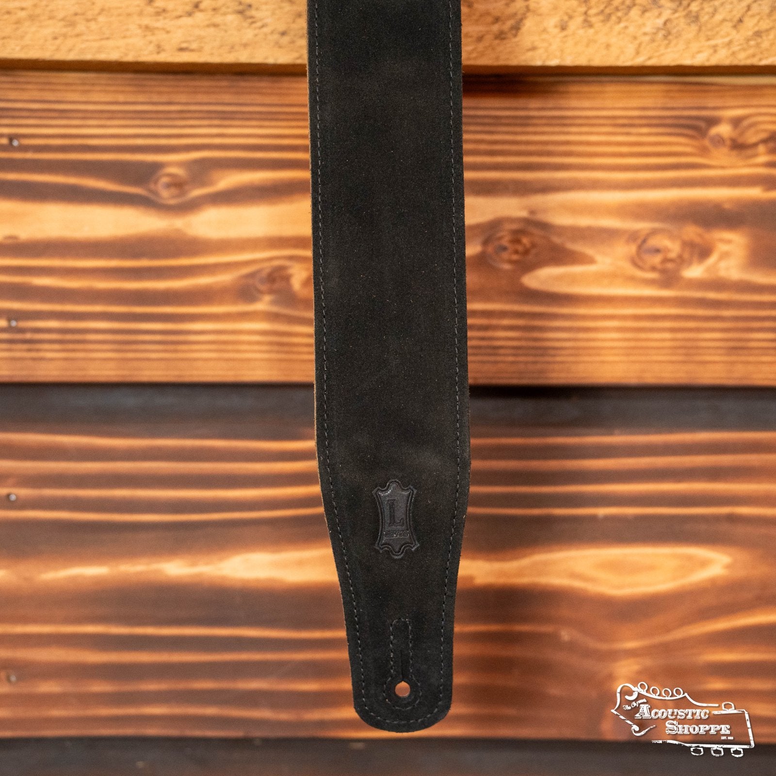 The Levy's MS26-BLK 2.5" suede guitar strap by Levy's Leather hangs vertically on a wooden wall, featuring a visible logo patch and detailed stitching. The warm, horizontal wood planks highlight the strap’s elegant, adjustable design.