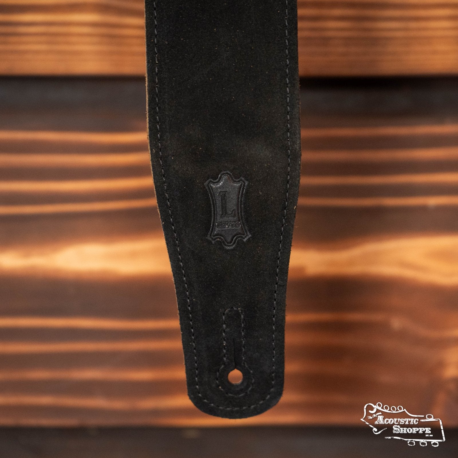 A close-up of the Levy's MS26-BLK 2.5" black suede guitar strap by Levy's Leather, featuring a stitched edge and embossed L logo, hangs against a wooden background. The Acoustic Shoppe logo is in the lower right corner.
