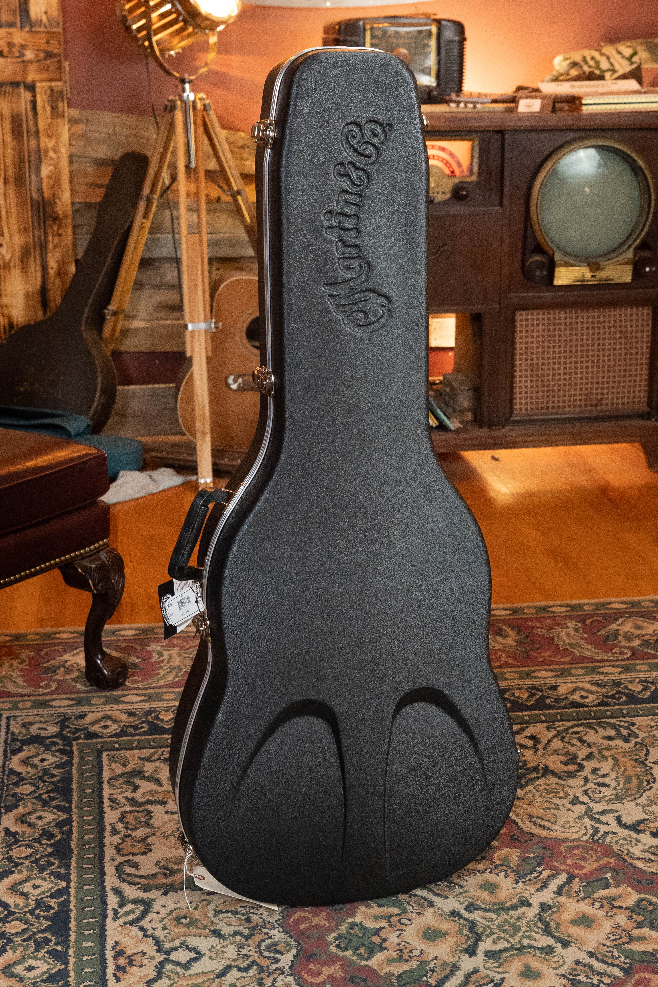 A black Martin & Co. guitar case, possibly holding a Martin Custom Shop HD-28 Style Spruce/Wild Grain Rosewood Custom "D" Dreadnought Acoustic Guitar #7410, stands on a patterned rug in a cozy, vintage-themed room with musical instruments and retro decor.