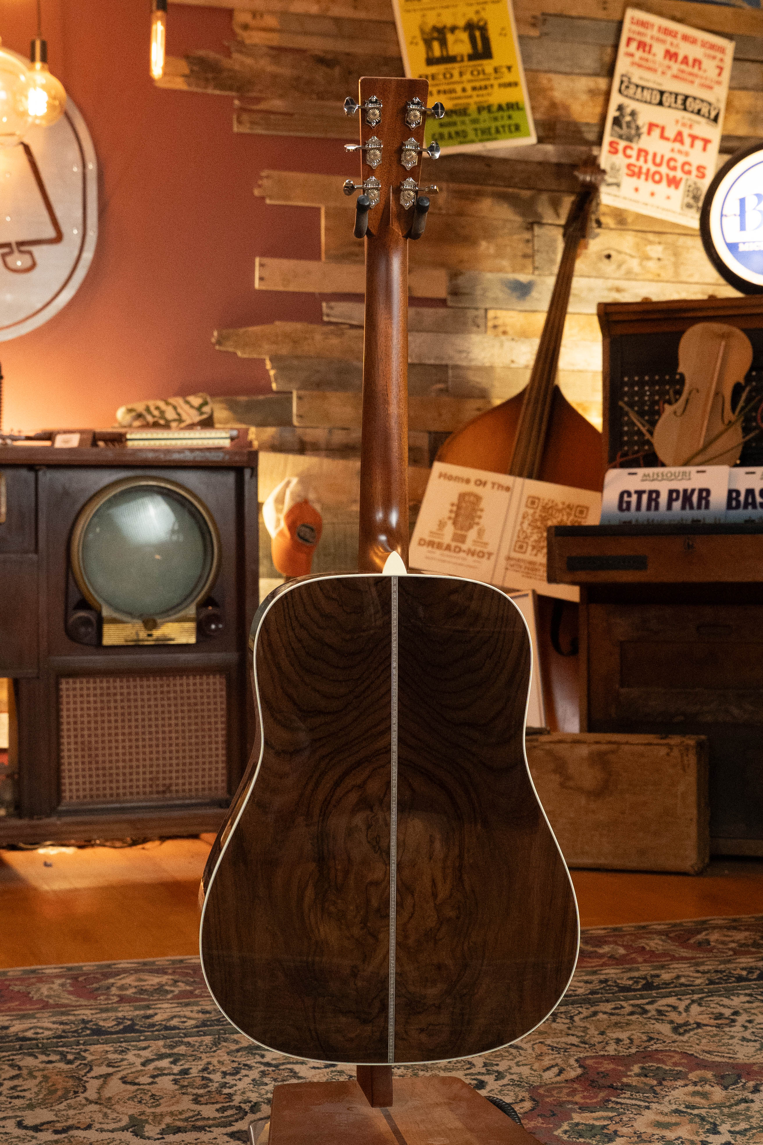 The Martin & Co. Custom Shop HD-28 Style Spruce/Wild Grain Rosewood Custom "D" Dreadnought Acoustic Guitar #7410 is shown back-first in a warm, vintage-inspired music room filled with instruments, posters, and cozy lighting.