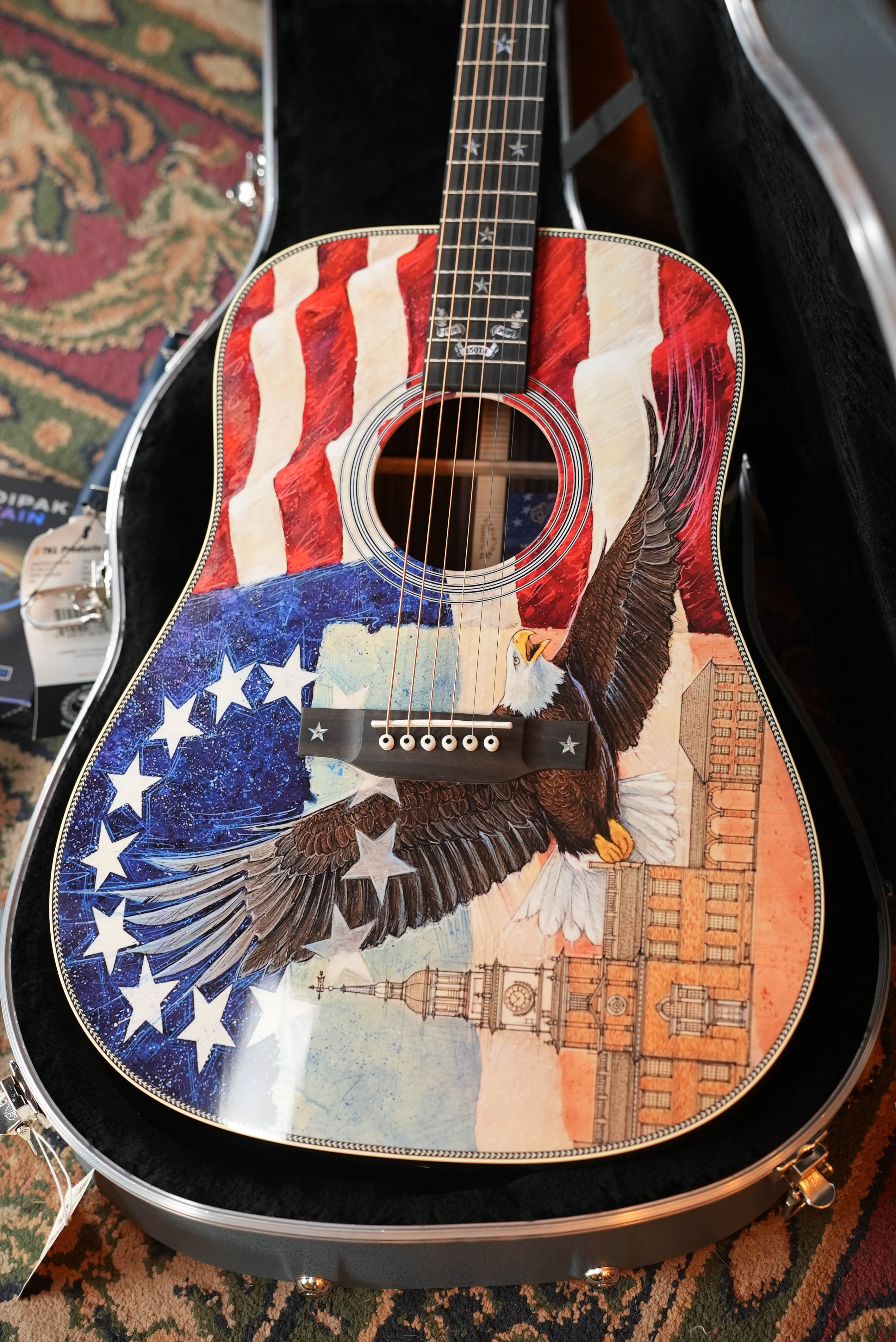 The Martin & Co. HD-28 Semiquincentennial Limited Edition Custom Ink Design Acoustic Guitar #7873, featuring an American flag, bald eagle, stars, and historic buildings, rests in an open black case on a patterned carpet.