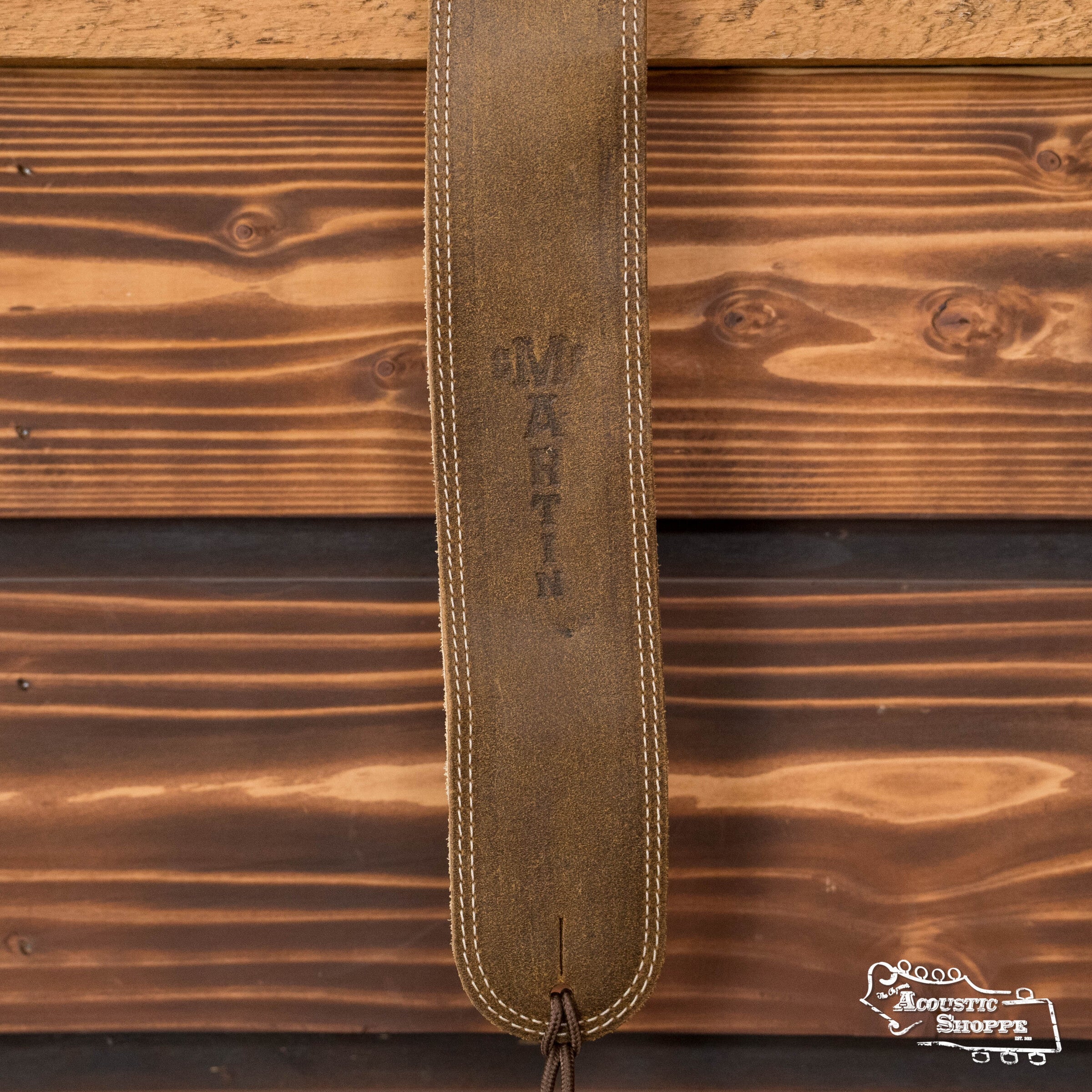 A close-up shows the Martin & Co. 2.5" Light Brown Distressed Ball-Glove Leather Guitar Strap, embossed with "MARTIN," hanging against a wooden wall. Available as part of The Acoustic Shoppe’s acoustic instrument accessories lineup.
