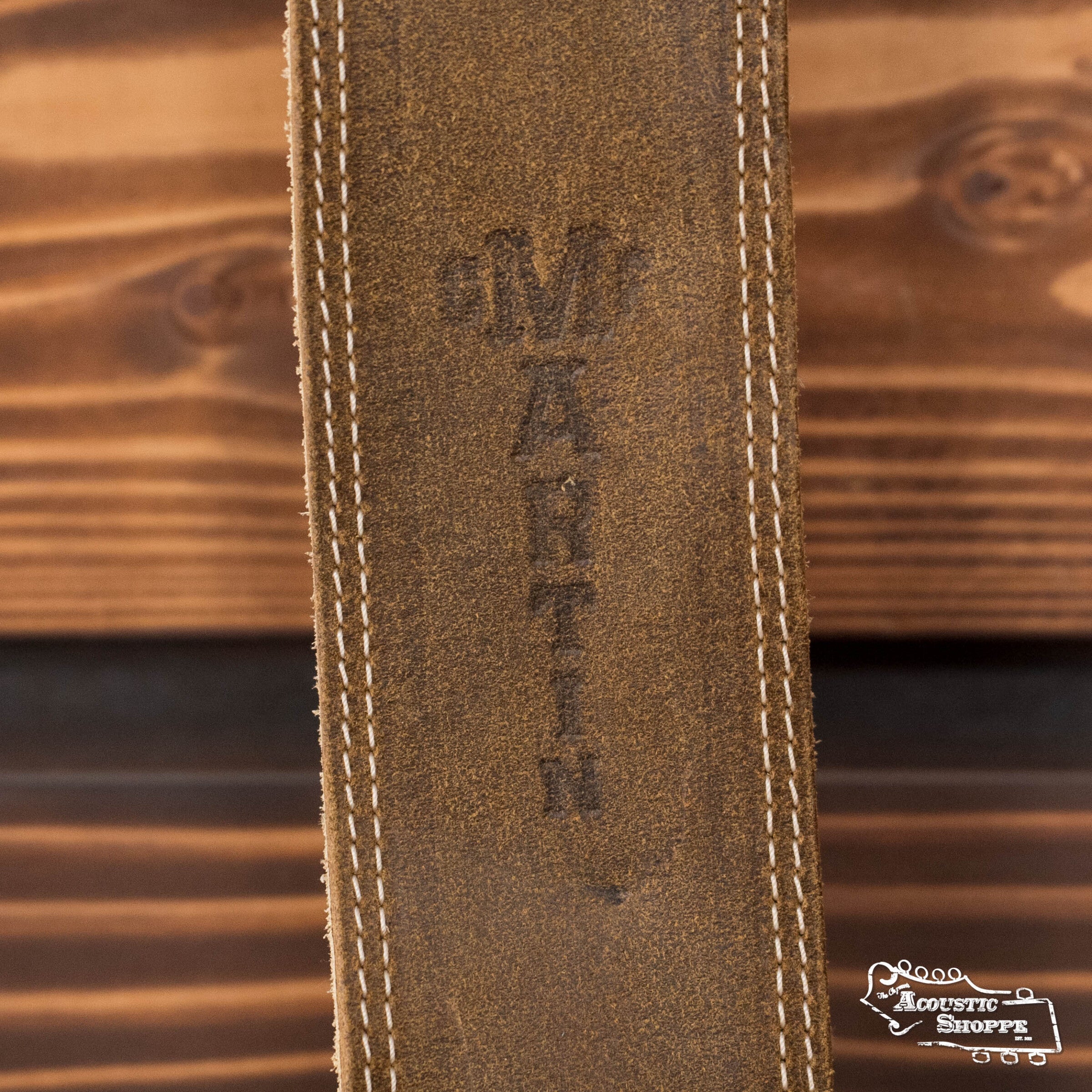 Close-up of the Martin & Co. 2.5" Light Brown Distressed Ball-Glove Leather Guitar Strap with white stitching and vertical embossing, shown on wood with The Acoustic Shoppe logo in the bottom right corner. Perfect for acoustic accessories.