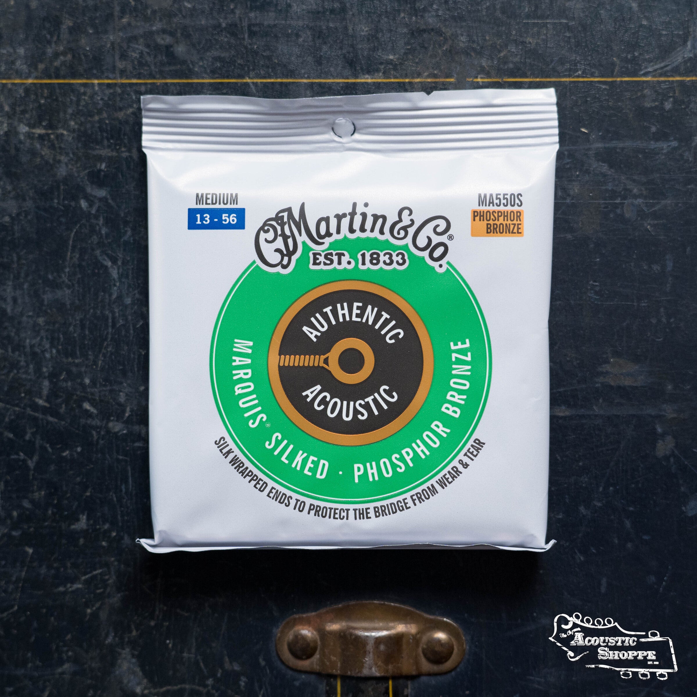 A pack of Martin & Co. Authentic Medium Marquis Silked Guitar Strings—Phosphor Bronze—rests on a dark surface with a metal latch beneath, highlighting the signature silk wrap of these Martin & Co. strings.