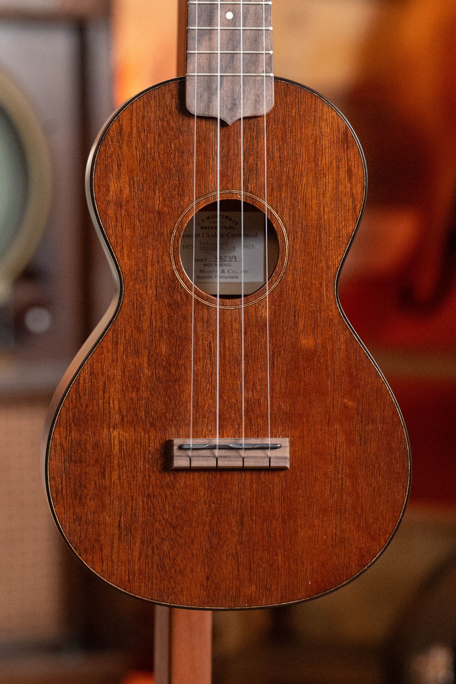 A close-up of the Martin & Co. Centennial Concert All Mahogany Ukulele #8239 highlights its mahogany body, strings, soundhole, and neck with a softly blurred background.