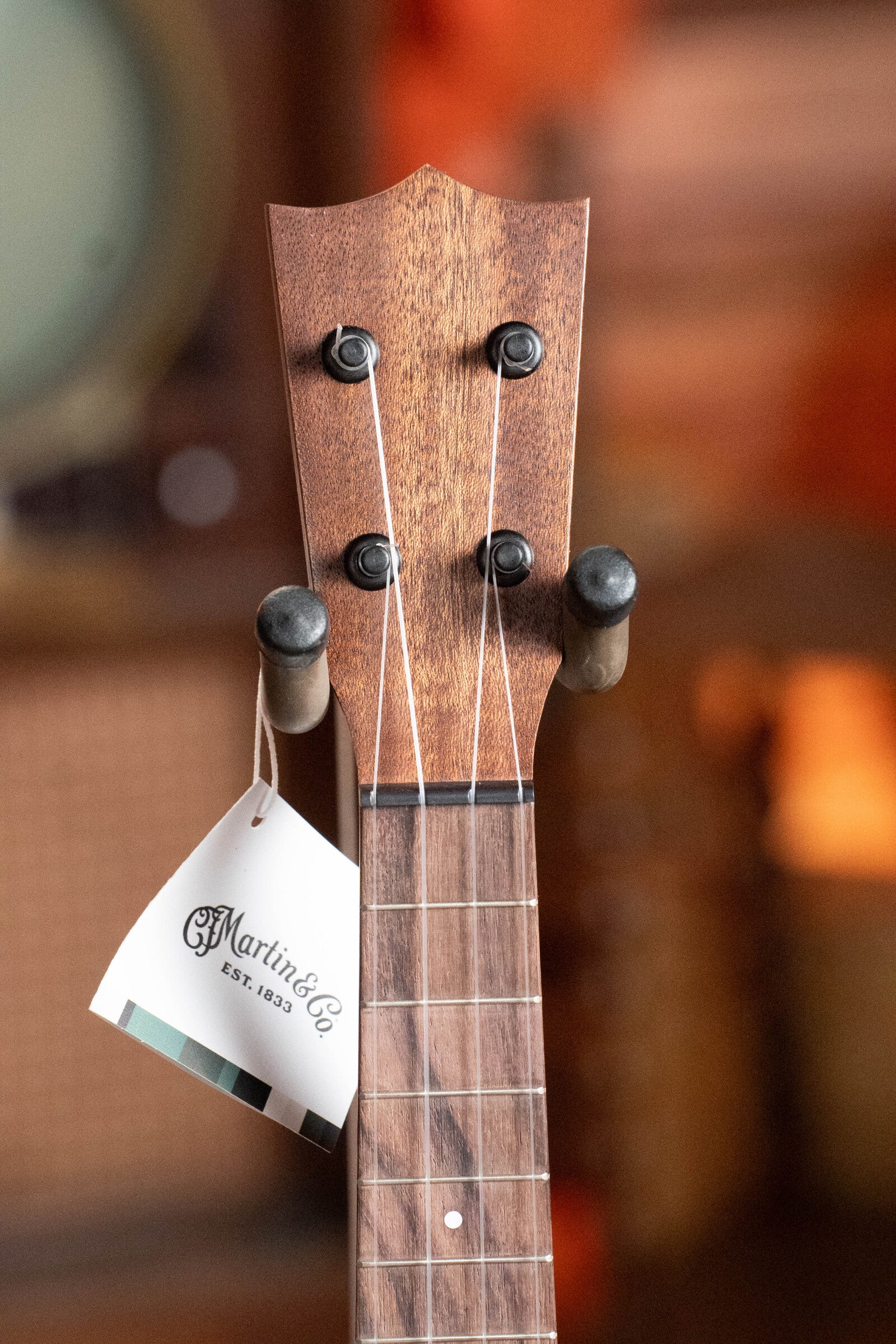 Close-up of the Martin & Co. Centennial Concert All Mahogany Ukulele #8239 headstock, featuring four tuning pegs and a Martin & Co. tag, with a blurred background—ideal for fans of all-mahogany acoustic ukuleles.
