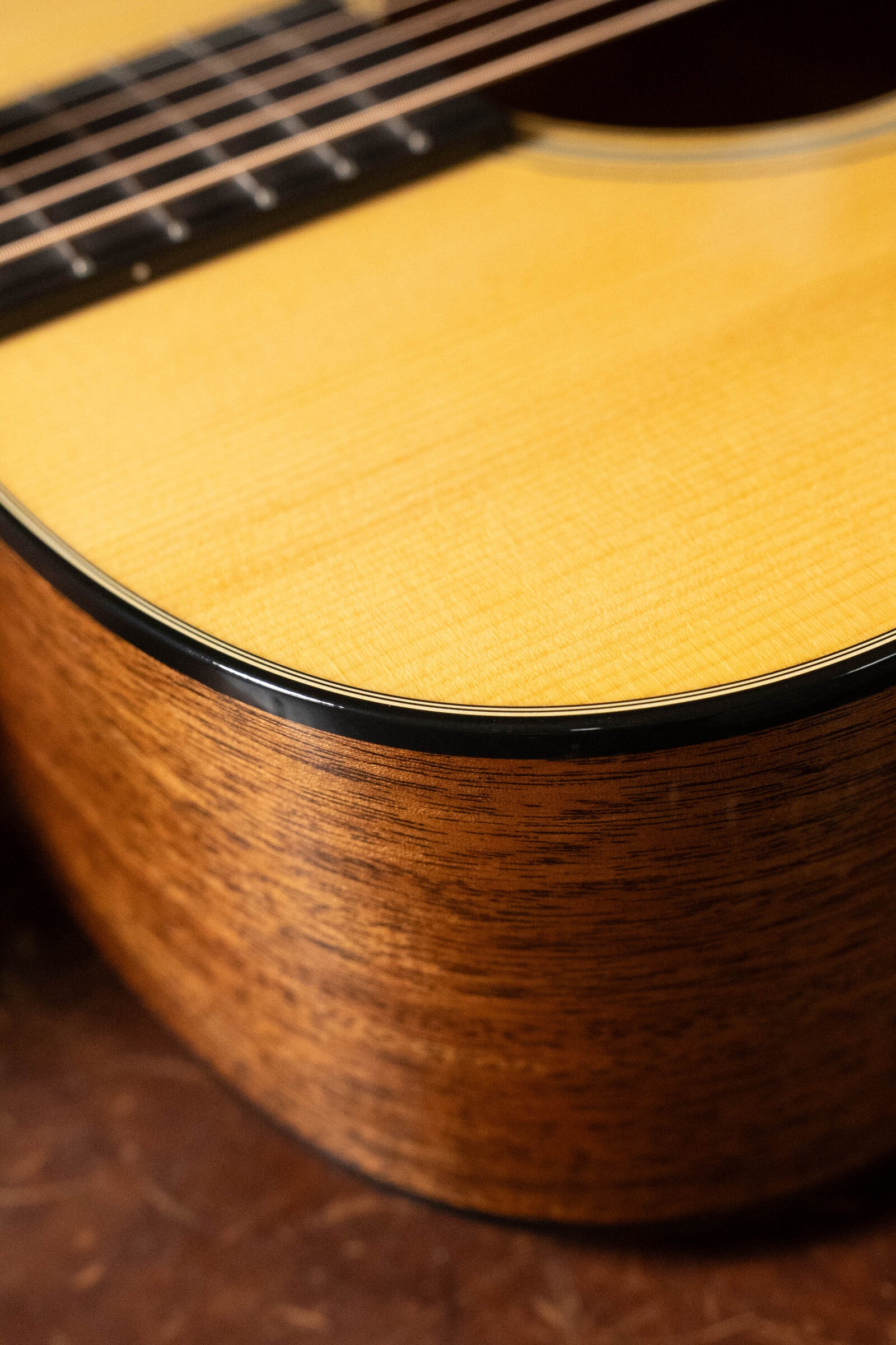 Close-up of a Martin & Co. Custom Shop D-18V Adirondack/Mahogany Dreadnought Acoustic Guitar #6905, showcasing its glossy Adirondack spruce top, smooth grain, bridge, and dark brown mahogany sides on a textured surface.