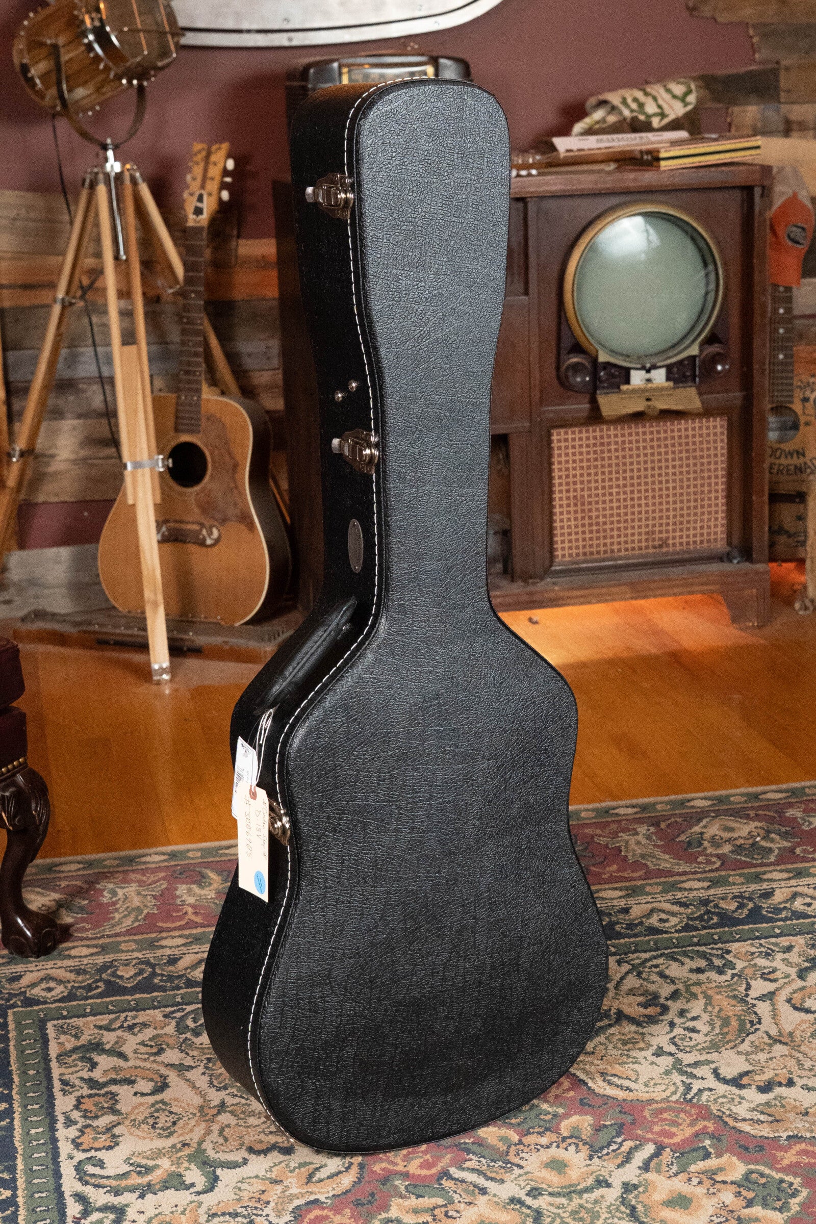 A closed black guitar case stands on a patterned rug in a cozy room, with a Martin & Co. Custom Shop D-18V Adirondack/Mahogany Dreadnought Acoustic Guitar #6905, an old TV, and wooden furniture in the background.