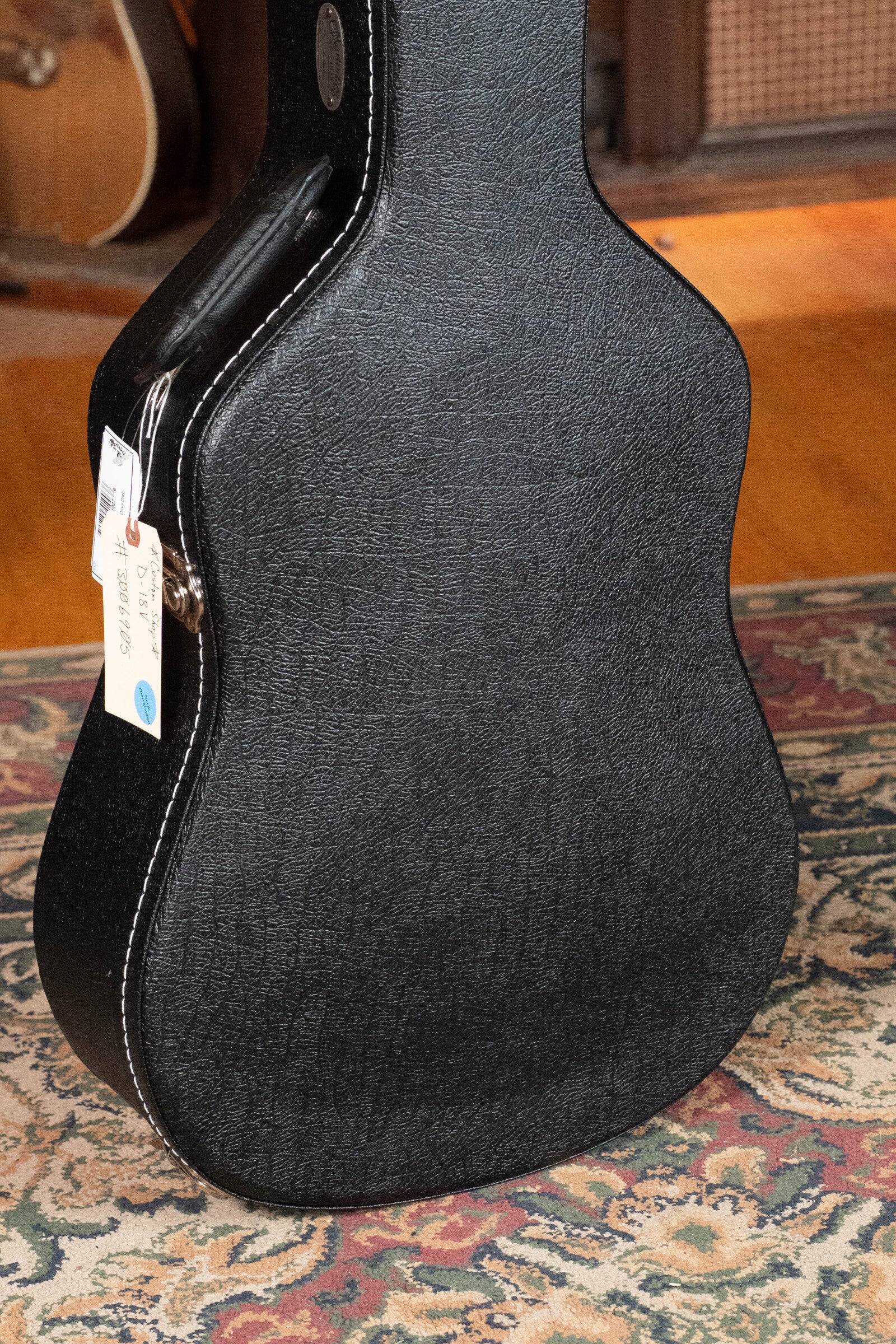 A black textured guitar case with a handle and tags stands upright on a patterned rug, with part of a Martin & Co. Custom Shop D-18V Adirondack/Mahogany Dreadnought Acoustic Guitar #6905 and speaker visible in the background.