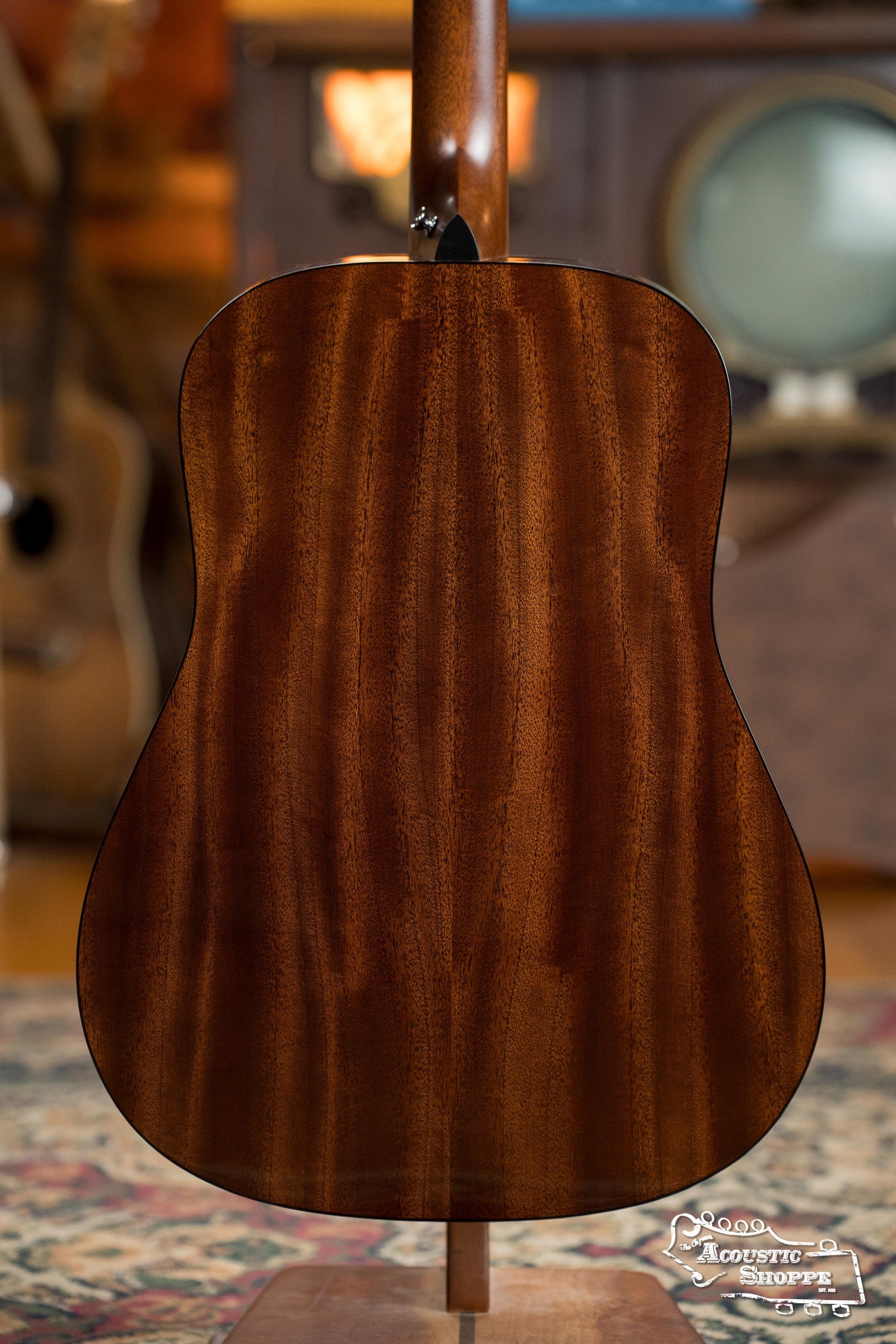 The image displays the back of a Martin & Co. DE Retro Plus Torrefied Spruce/Mahogany Dreadnought Guitar w/Martin Electronics #0808, glossy brown wood finish, upright on a stand with blurred musical instruments and vintage decor in the background.