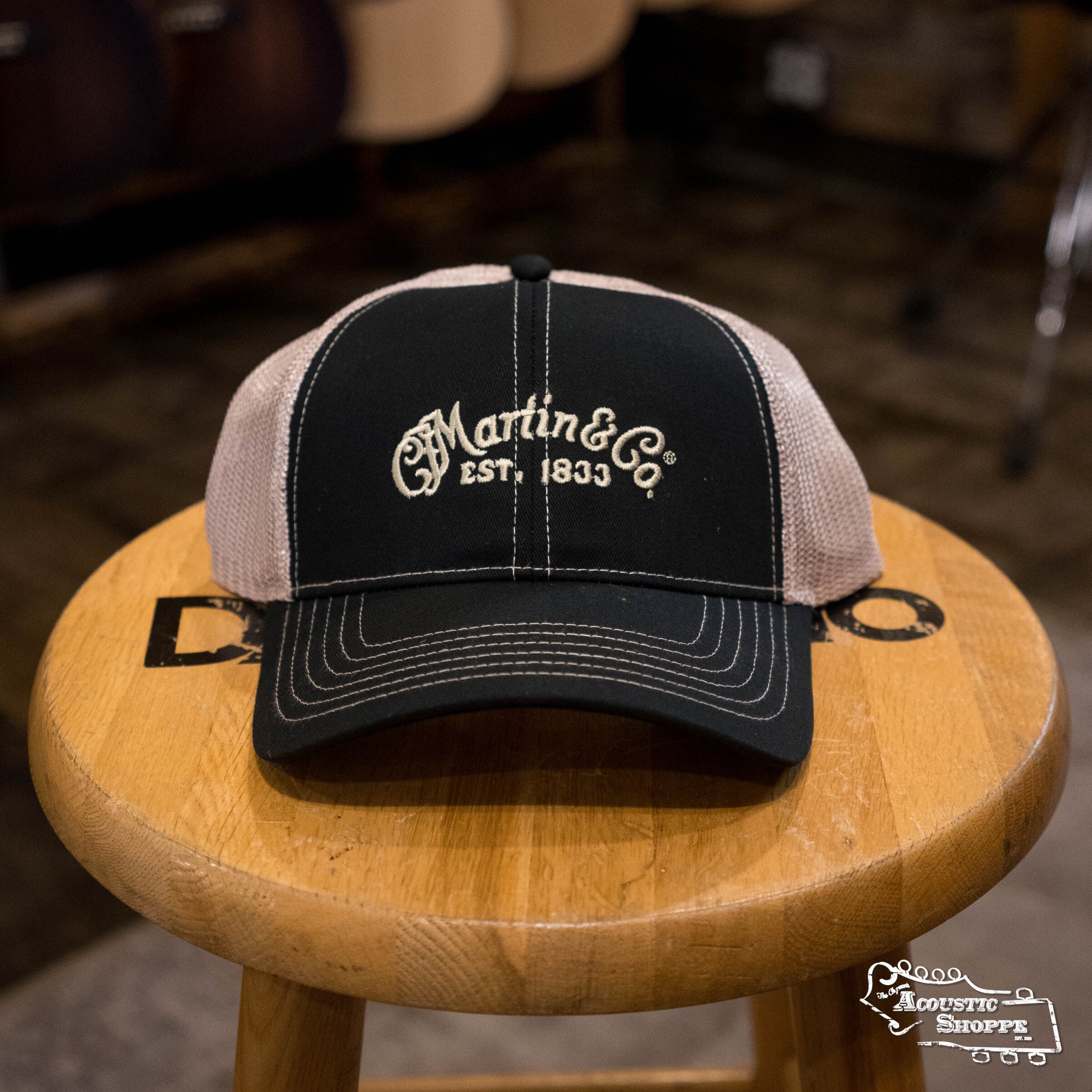 The Martin & Co. Martin Logo Mesh Trucker Hat - Black with an adjustable strap is displayed on a wooden stool with The Acoustic Shoppe logo, in a store setting with blurred guitars in the background.
