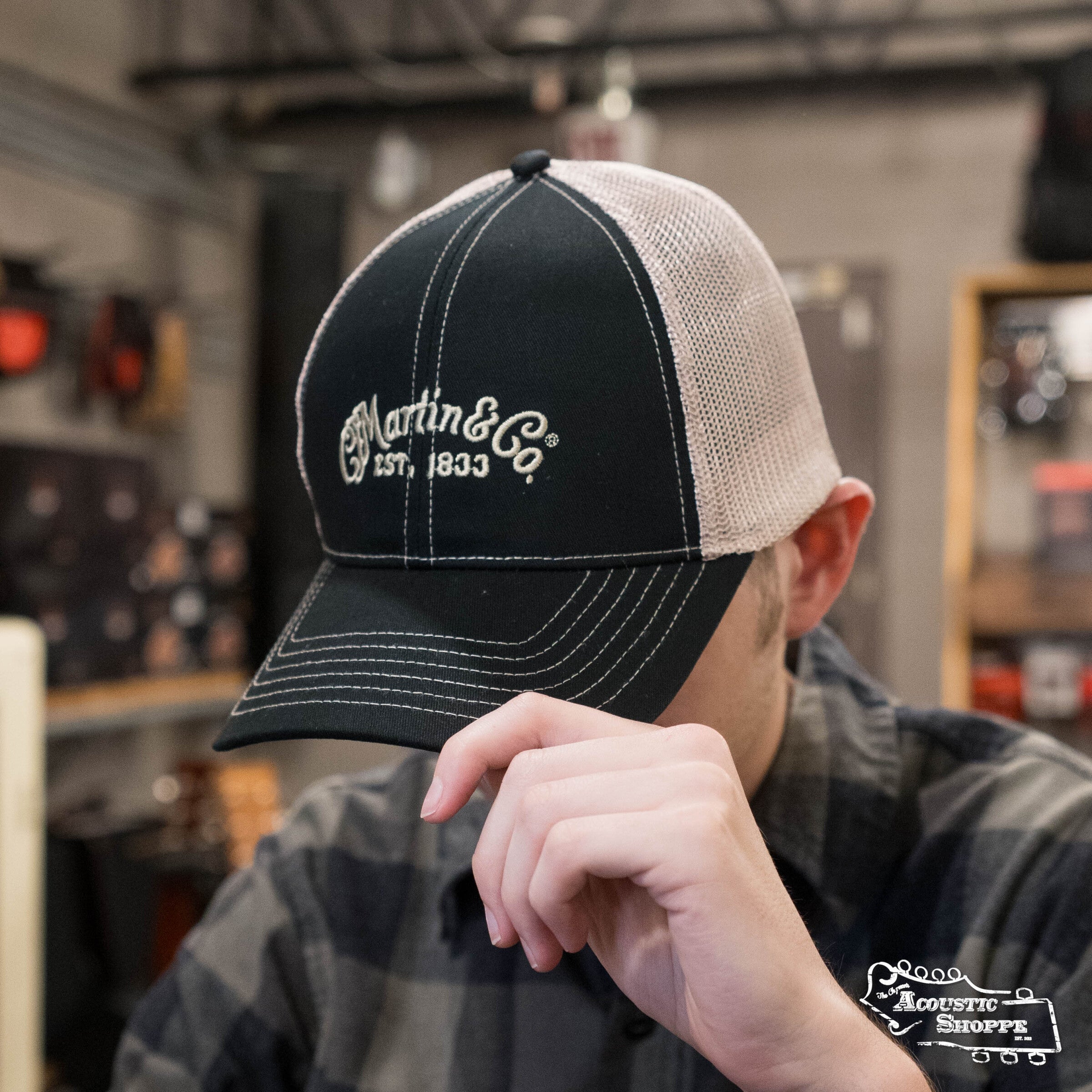 A person tilts the brim of a Martin & Co. Martin Logo Mesh Trucker Hat - Black, featuring a black and white design with “EST. 1833” embroidery. The adjustable strap shows at the back, blurred musical instruments in the background.