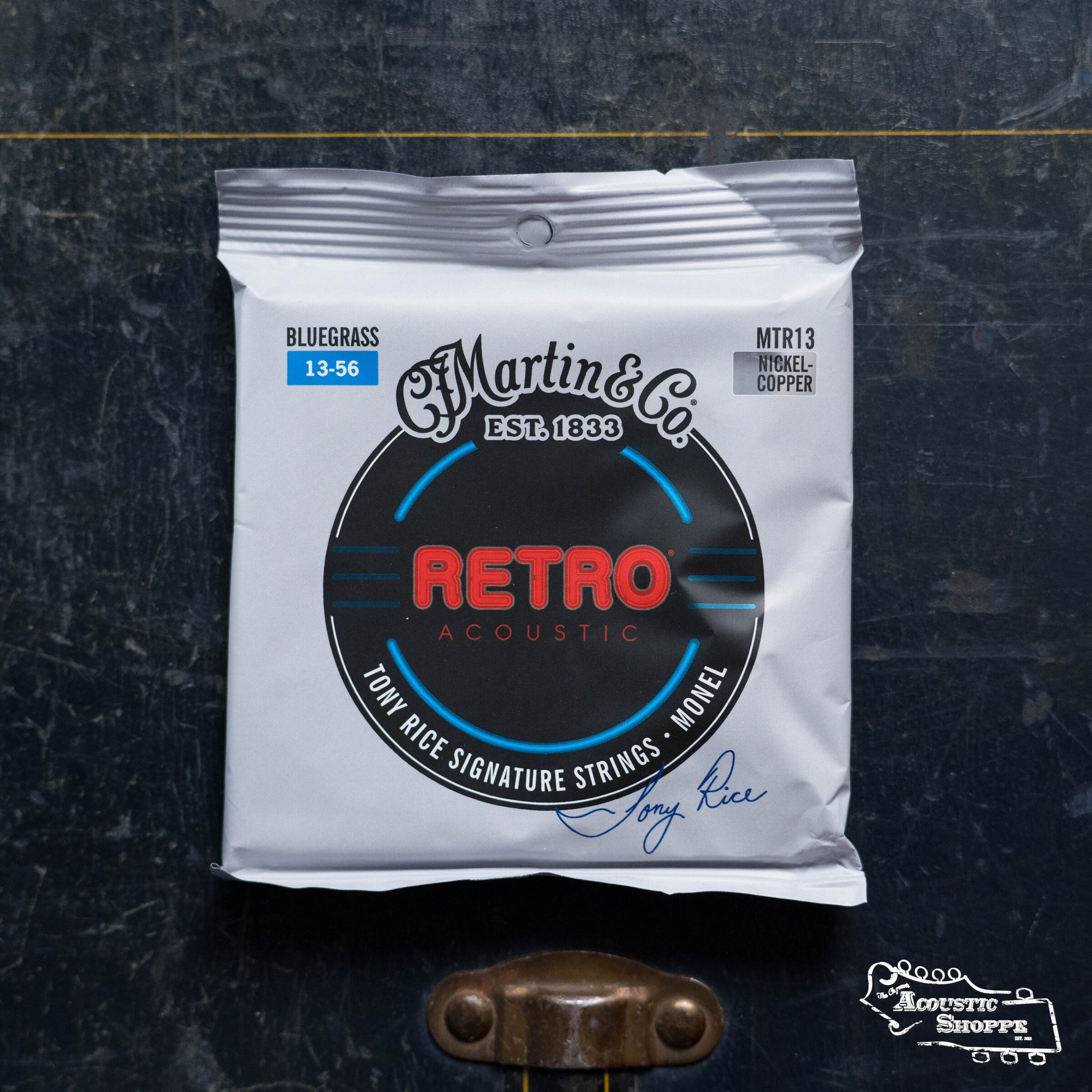 A pack of Martin & Co. Martin Retro Bluegrass-Tony Rice's Choice Acoustic Guitar Strings (41Y19MM13) rests on a dark, worn wooden surface with metal hardware and The Acoustic Shoppe logo.