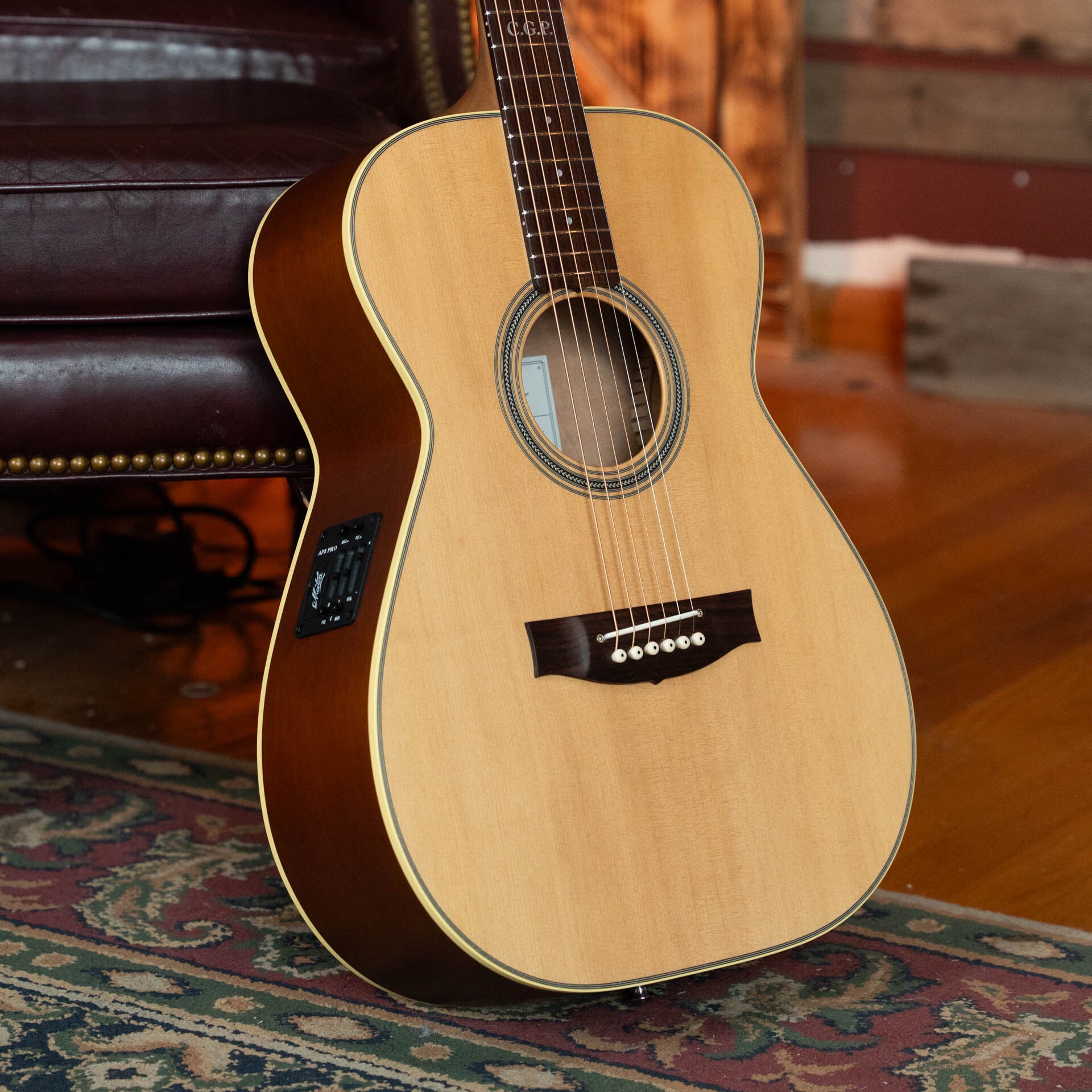 A Maton Guitars T.E. Personal Spruce/Queensland Maple Deep Body OO Guitar w/Electronics (#0085), with a natural finish, rests on a decorative rug by a brown leather chair. It features an AP5 PRO pickup and built-in electronic tuner panel.