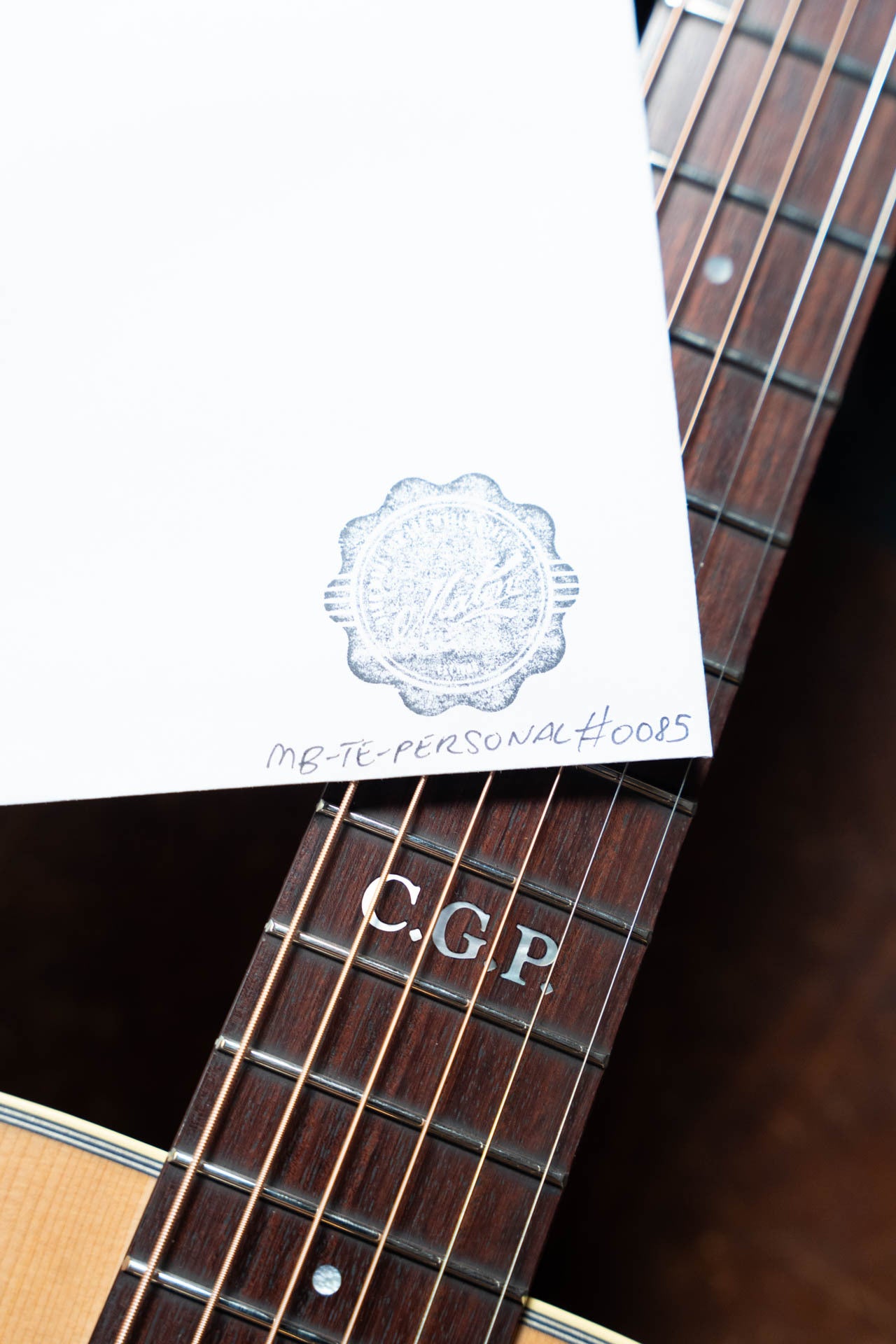 A white paper stamped MB-TE-PERSONAL#0085 rests on the fretboard of a Maton Guitars T.E. Personal Spruce/Queensland Maple Deep Body OO Guitar w/Electronics #0085, featuring C.G.P. inlays—a tribute to Tommy Emmanuels signature series.