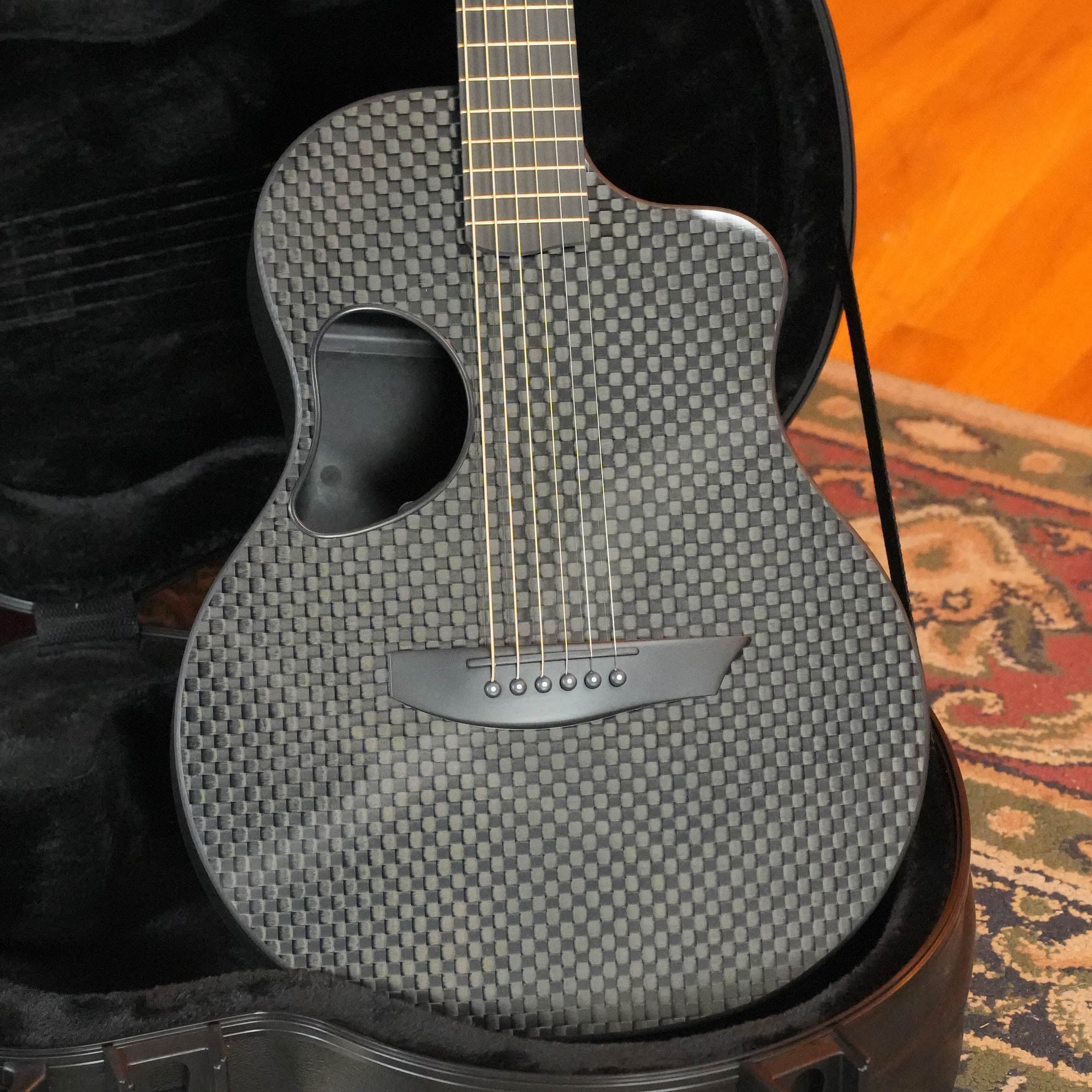 A close-up of the McPherson Guitars Carbon Fiber Touring Series Blackout Basketweave Guitar w/L.R. Baggs Element Pickup #0061, resting in its open case on a patterned rug—a top choice for touring musicians.