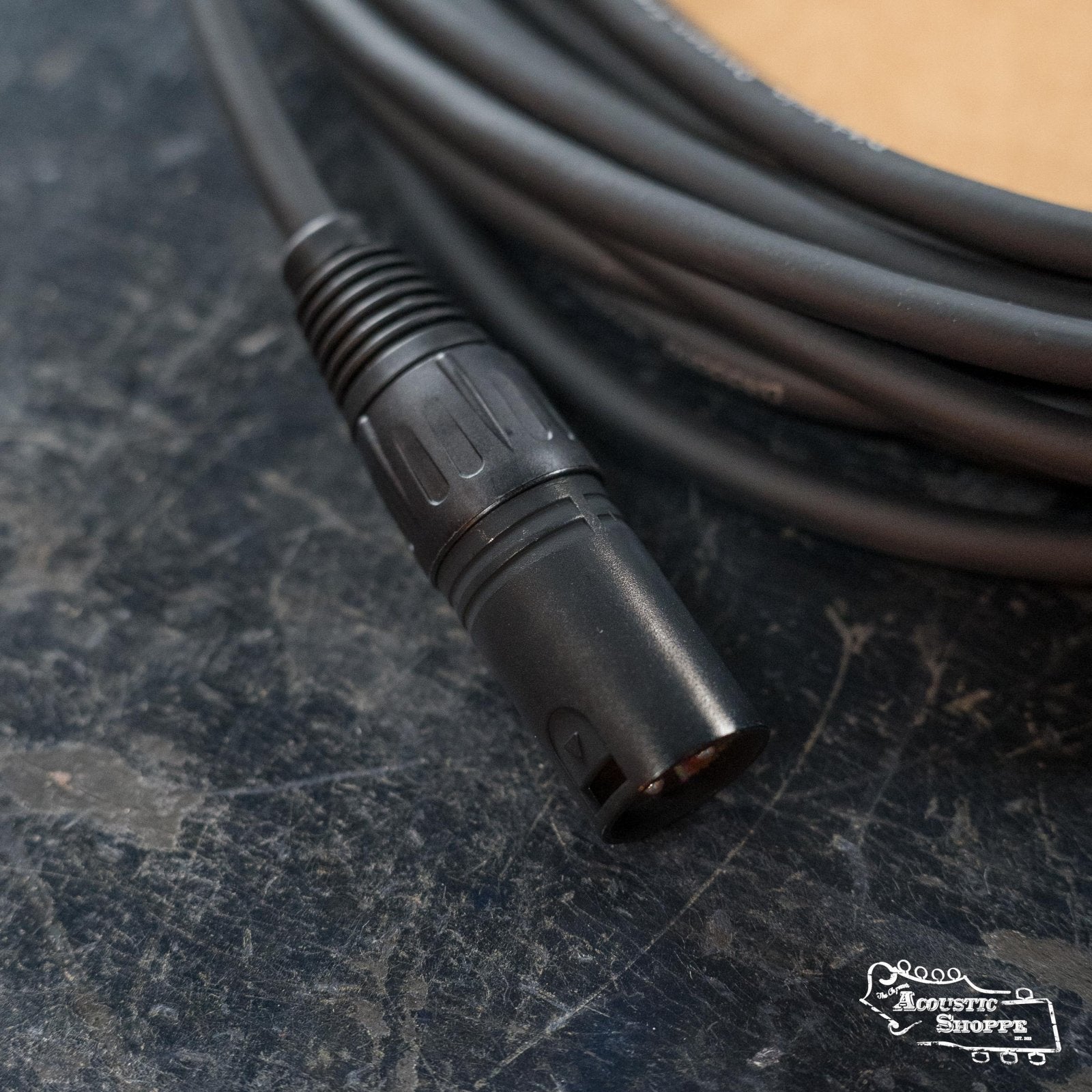 A close-up of a D'Addario Classic Series XLR Microphone Cable made with oxygen-free copper rests on a dark textured surface, connector in focus and coiled cable behind, with The Acoustic Shoppe logo in the bottom right corner.