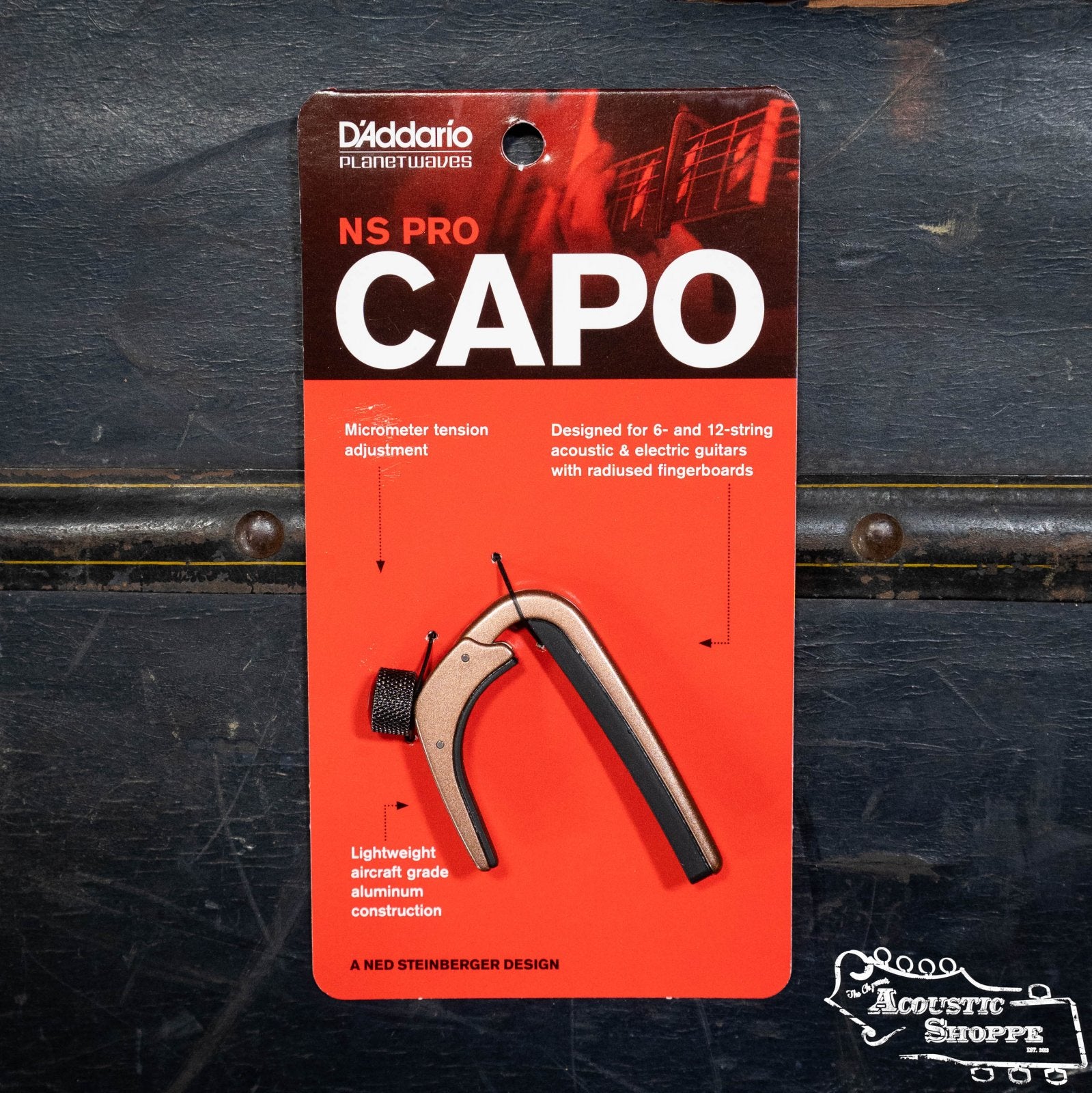 The D'Addario NS Capo Pro PW-CP-02 comes in red and black packaging with micrometer tension adjustment and lightweight aluminum features shown. The Acoustic Shoppe logo appears bottom right. Ideal capo for any guitaristโs setup.