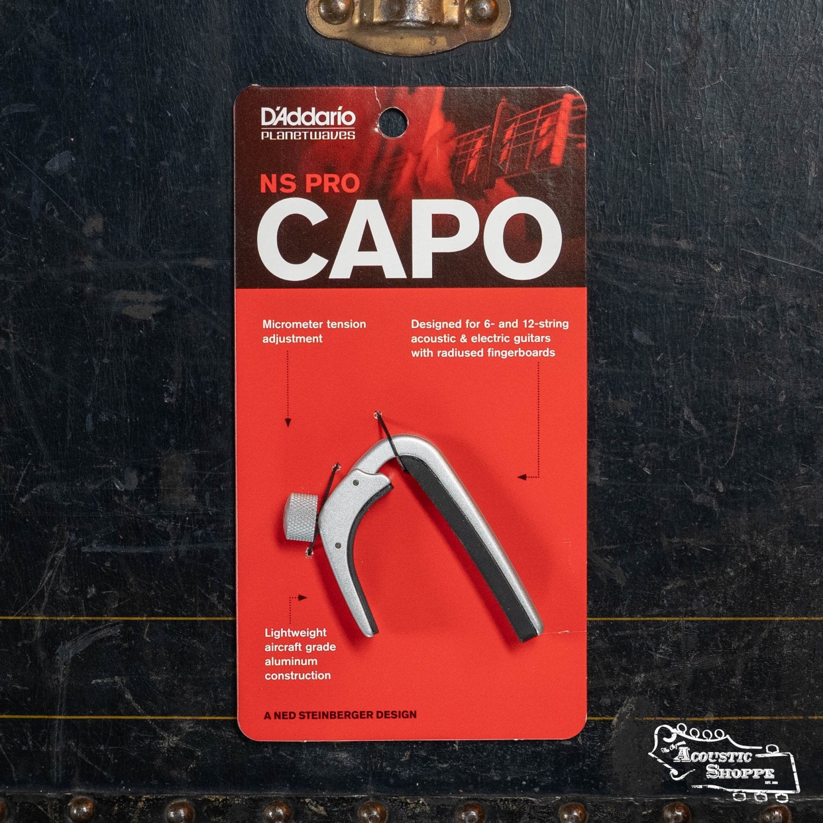 A D'Addario/Planet Waves NS Capo Pro PW-CP-02 in red and black packaging, showing product details and an image of the capo, is placed on a dark textured surface with the Acoustic Shoppe logo at the bottom right.