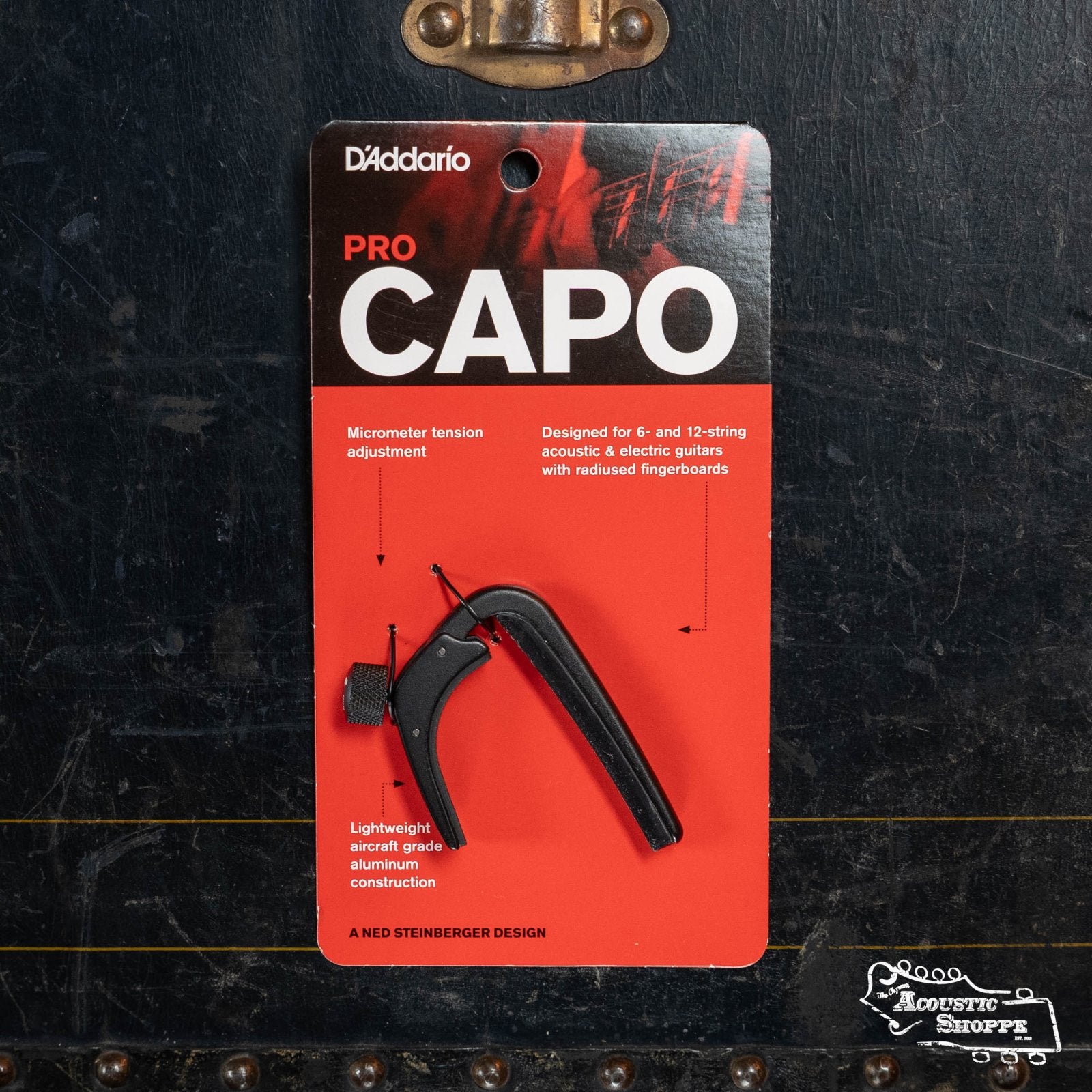 A D'Addario NS Capo Pro PW-CP-02 in black rests on its red and black packaging, highlighting micrometer tension adjustment and lightweight aluminum buildโa sleek, essential capo for guitarists from D'Addario.