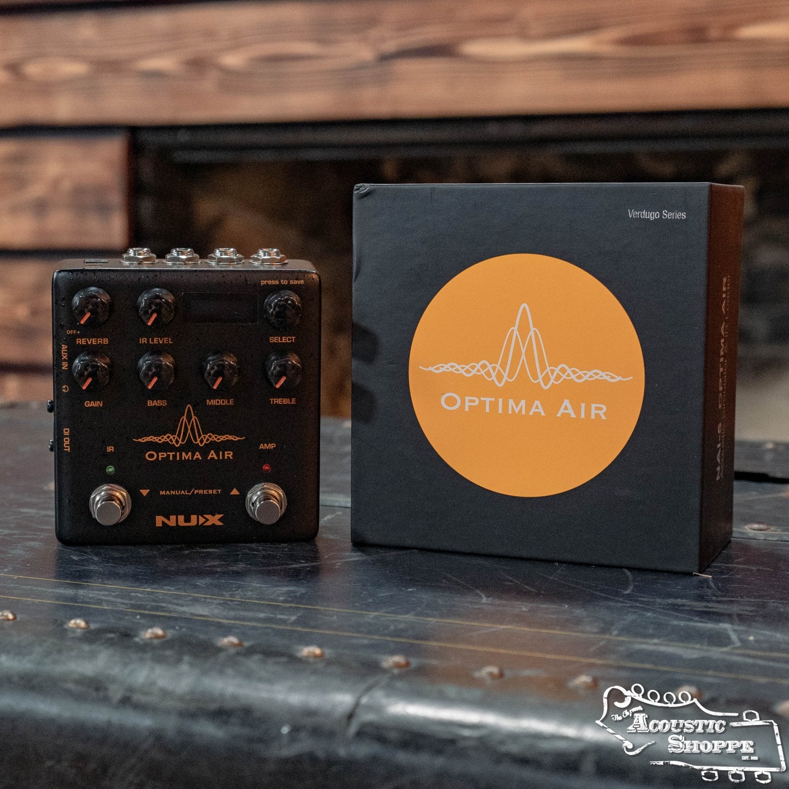A NUX NAI-5 Optima Air Acoustic Simulator & IR Loader from The Acoustic Shoppe sits by its black box with an orange logo on a black trunk, set against a blurred wooden background.
