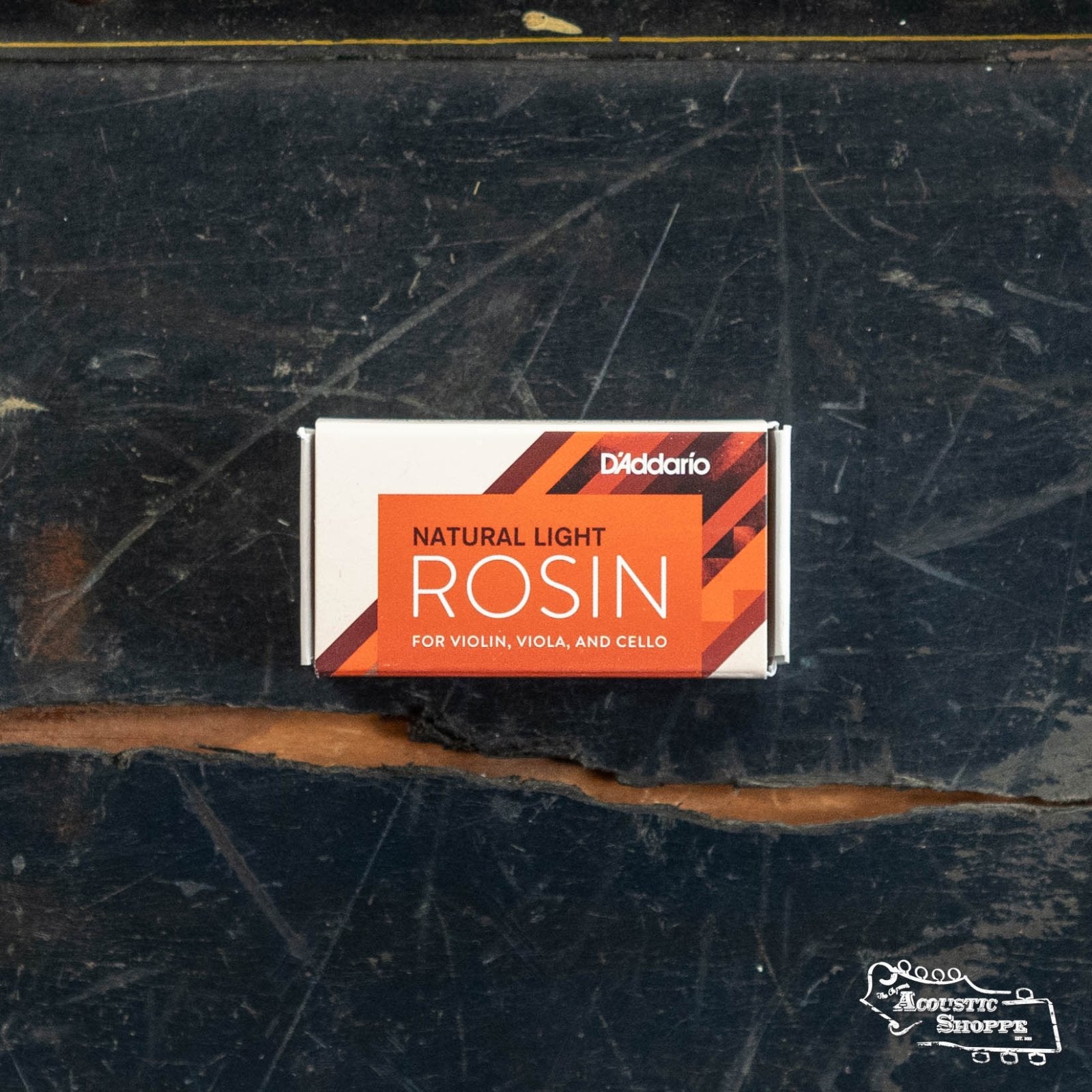 A box of DAddario Natural Rosin (VR200 VR300) for violin, viola, and cello by DAddario rests on a scratched, dark wooden surface. The box displays orange and red diagonal stripes.