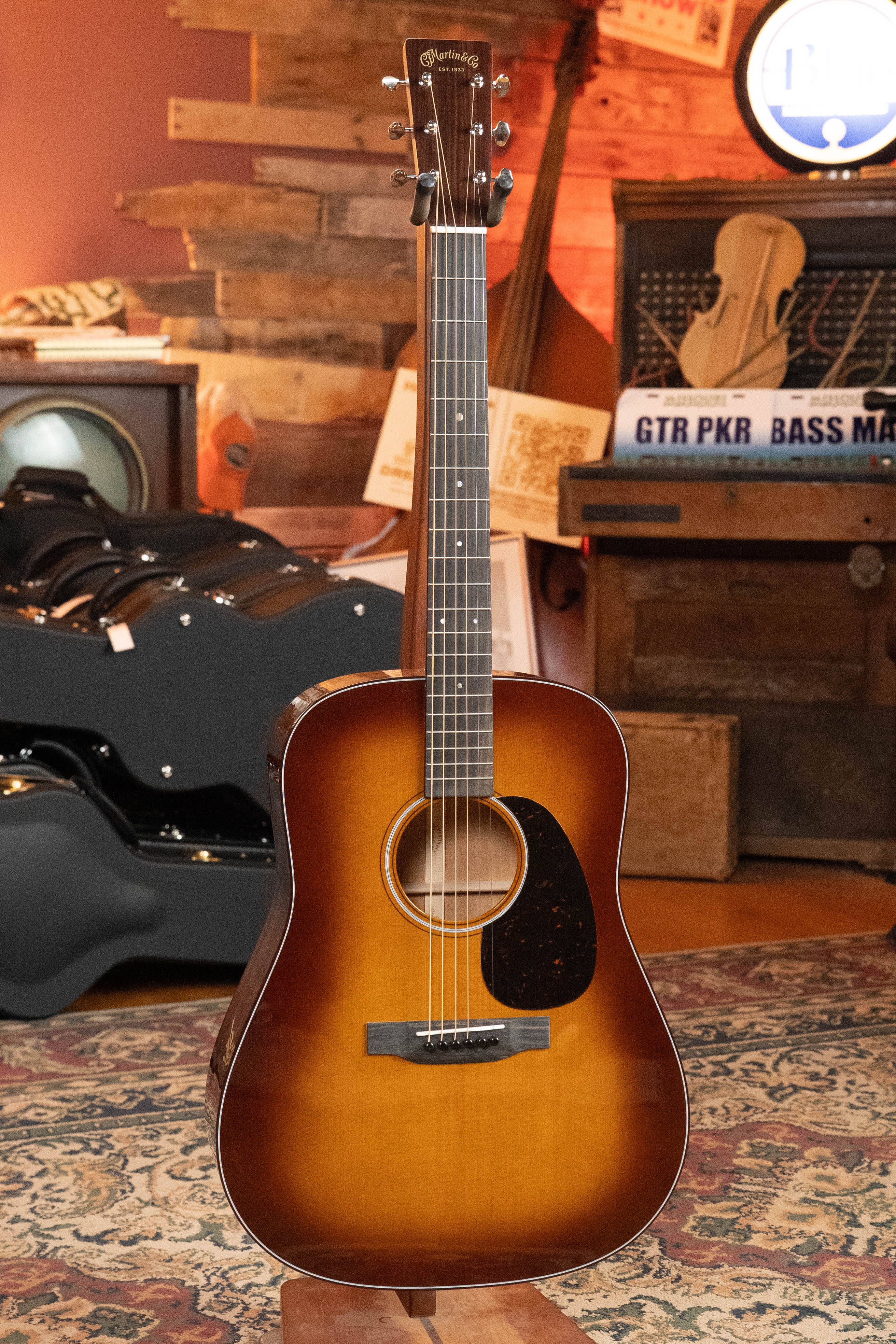 The *New 2025 Model* Martin D-18 1933 Gloss Ambertone Spruce/Mahogany Dreadnought Acoustic Guitar #6784 by Martin & Co. stands upright on a patterned rug in a cozy, warmly lit room with wooden decor and music equipment in the background.