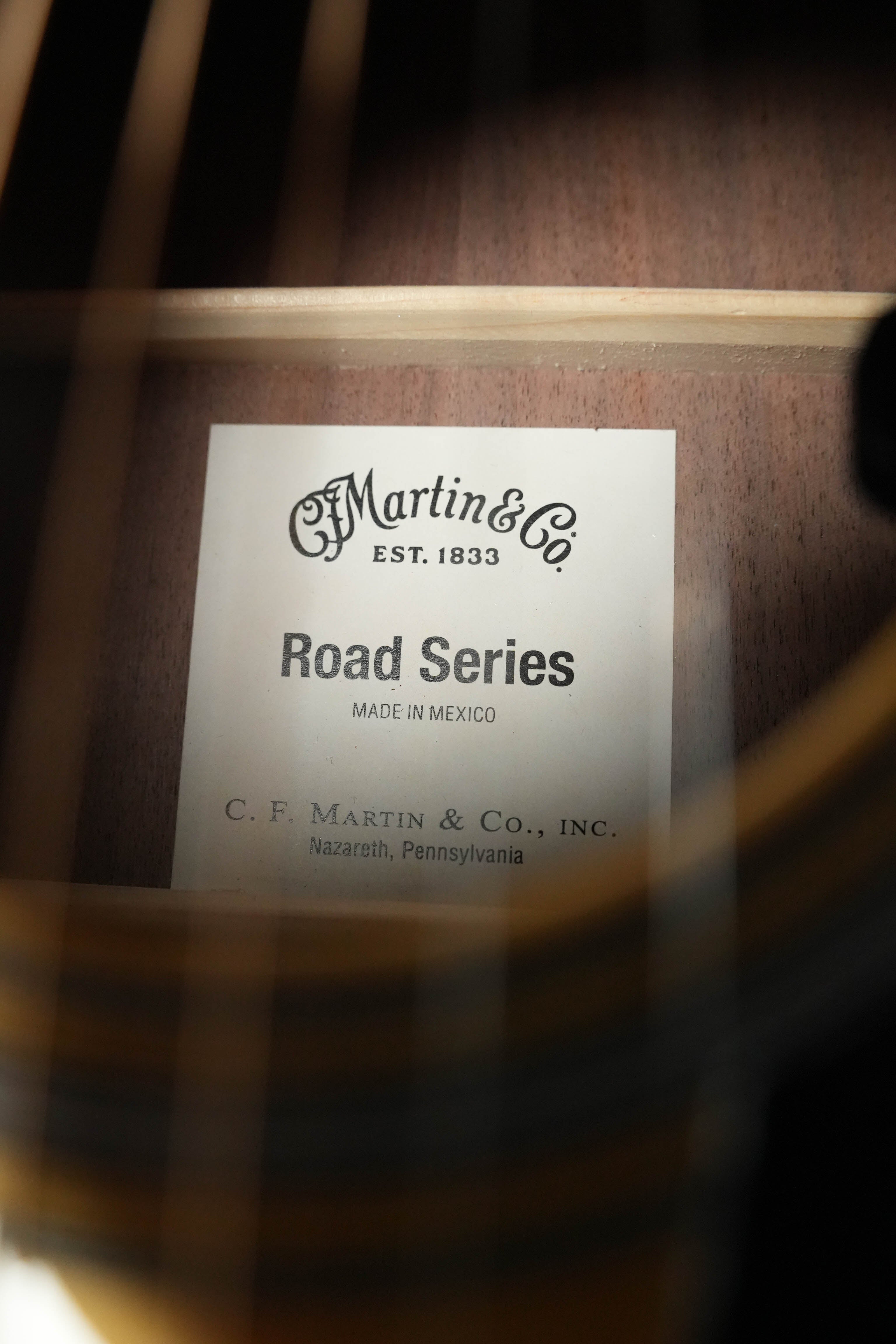 A close-up view through guitar strings reveals a label inside the Martin & Co. *New 2026 Model* D-13E Retro Road Series dreadnought, highlighting its quality craftsmanship and advanced Martin electronics.