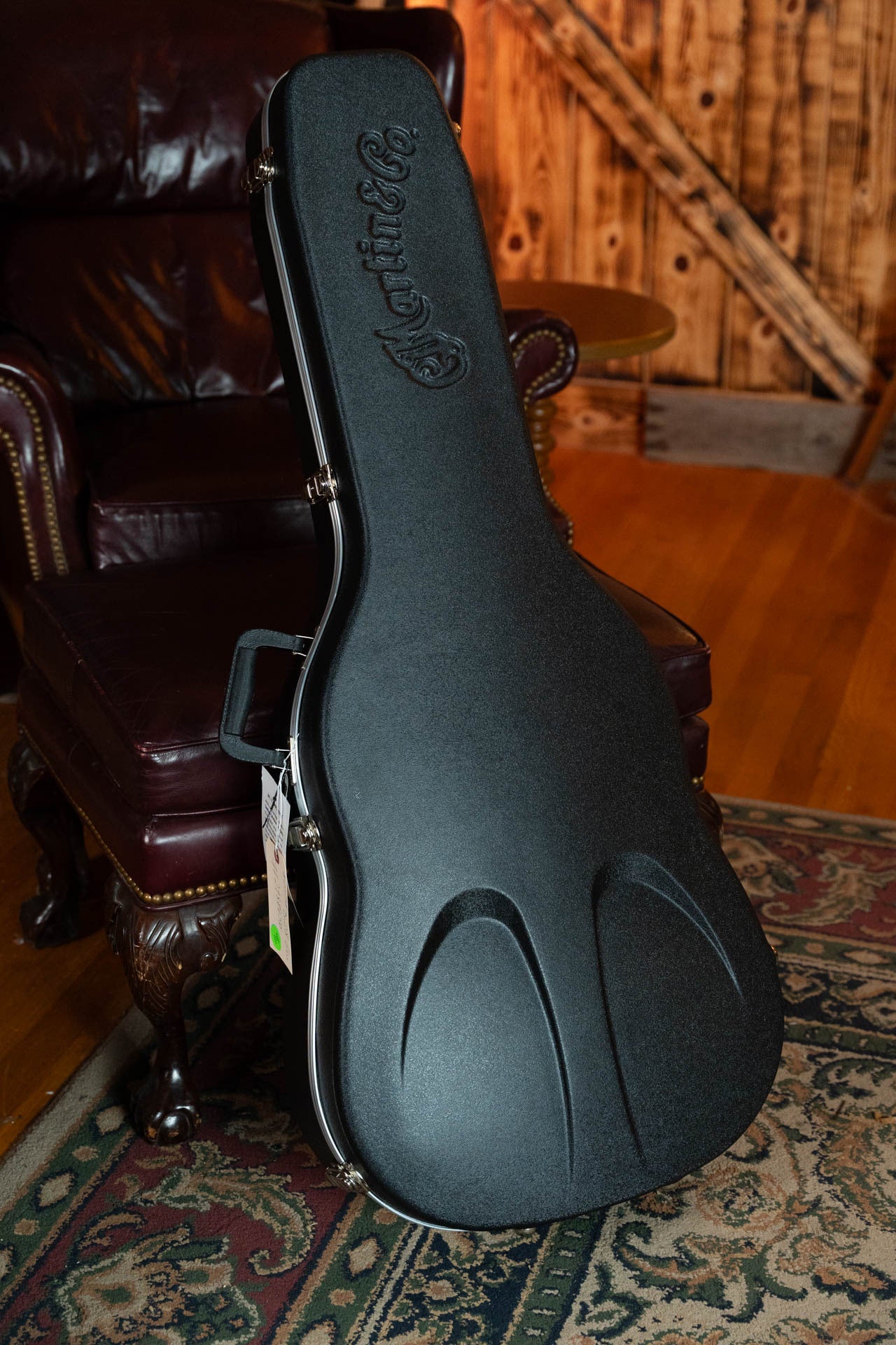 A black hard guitar case with Martin & Co. embossed on the lid rests against a leather armchair—ideal for your *New 2025 Model* Martin D-28 Gloss Spruce/Indian Rosewood Dreadnought Acoustic Guitar #0650.