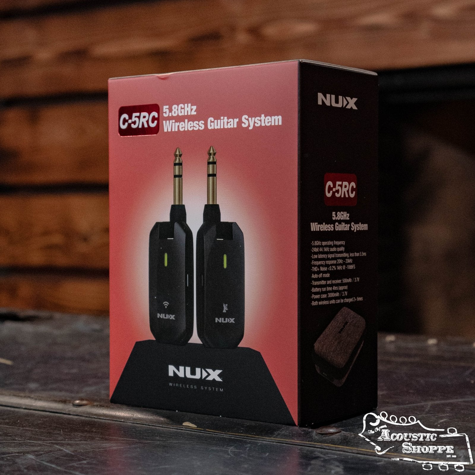 A boxed NUX C-5RC 5.8GHz Wireless Guitar System for Acoustic or Electric Guitar (Black) by NUX is shown on a wooden surface, with images of the transmitter and receiver on the front and a rustic wooden wall behind.